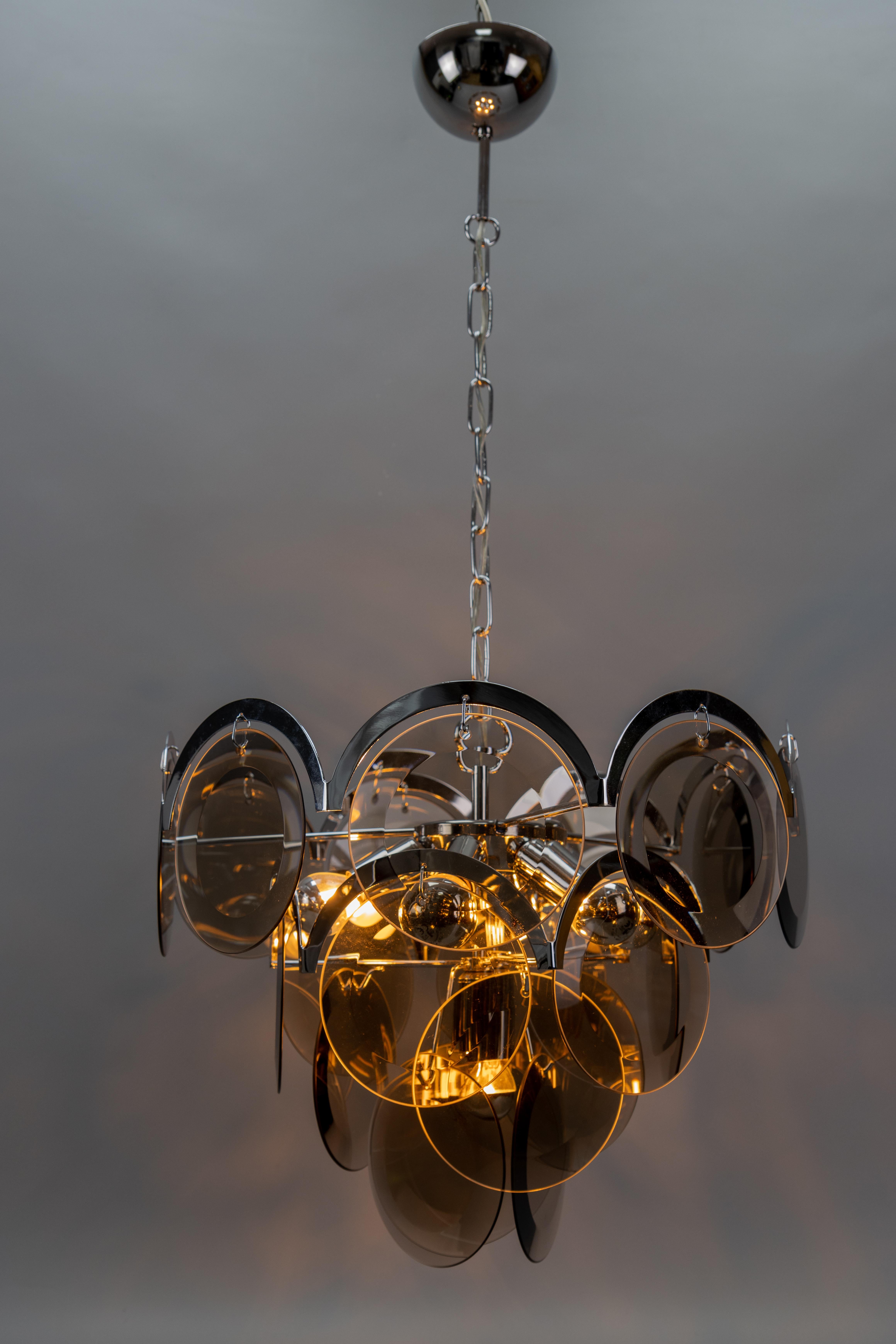 Lampadario a sospensione a sette luci in vetro fumé e metallo cromato, Italia, anni '70 circa, attribuito a Gino Vistosi.
Questo splendido lampadario in stile Mid-Century Modern presenta una struttura in metallo cromato a tre livelli ornata da 21