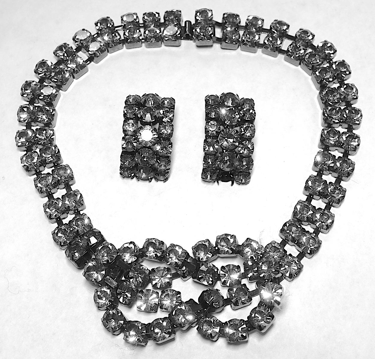 This stunning necklace and earring set is designed with smoky colored crystals with an encircled bow in the center. The necklace measures 17” and bow is 1-1/2” and is set with a Japanned finish.  The matching earrings are pierced and measure 1-1/4”