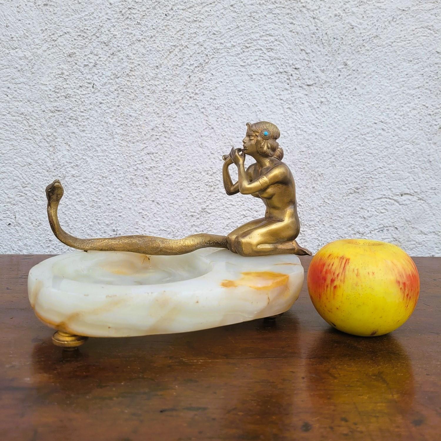 Incantevole centrotavola in bronzo patinato che rappresenta una giovane donna inginocchiata che suona il flauto davanti a un serpente.
L'incantatore di serpenti e il serpente sono fissati su una base in onice

Scultura nello stile delle creazioni di