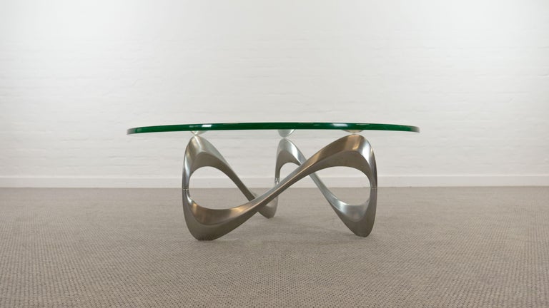 Mid-century coffee table - Sofa Tabl, designed by Knut Hesterberg, manufactured by Ronald Schmitt, Germany. Massive freeform aluminium base with the appearance of an infinity ribbon. Round glass-top. Clear glass, with green edge. Glass-thickness: