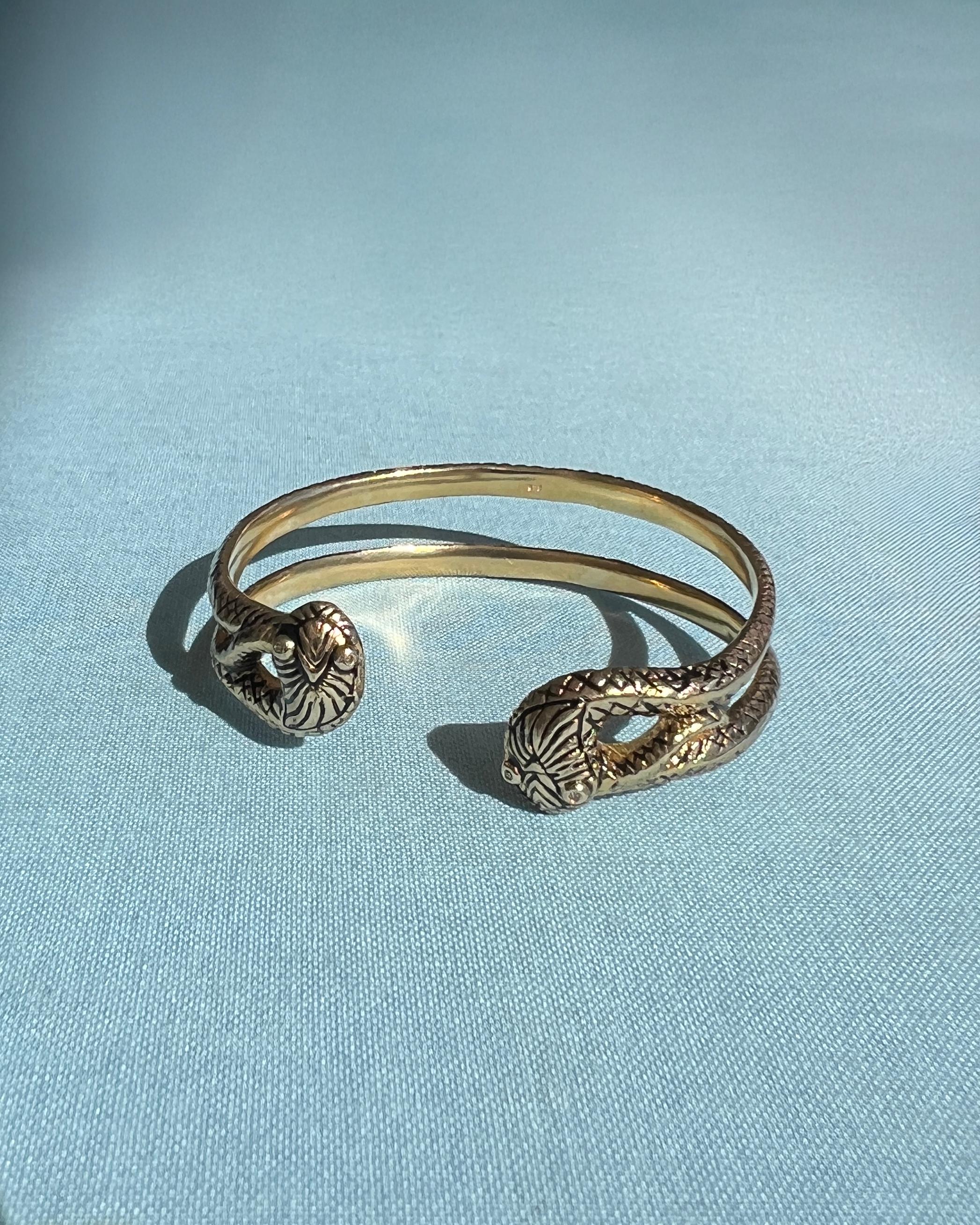 Manschettenarmband mit zwei Schlangen im viktorianischen Stil von J DAUPHIN aus Sterlingsilber mit 14-karätiger Vergoldung und handgemalter schwarzer Emaille für einen antiken Look.
Das Design zeigt zwei einander zugewandte Schlangen, die jeweils