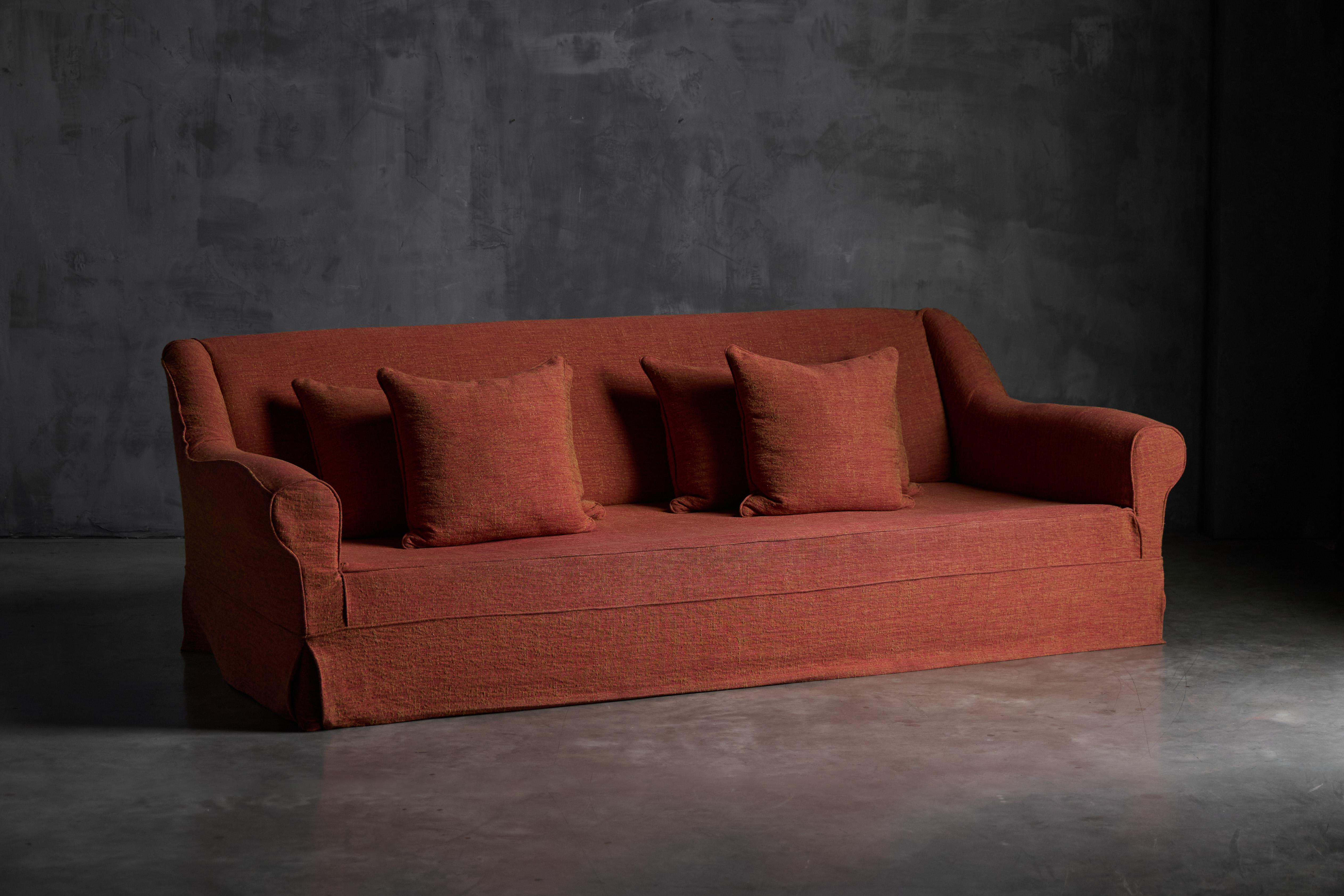 This Axel Vervoordt sofa is upholstered in a warm, orange rust-toned linen blend, the fabric has a natural, textured look that feels both cosy and refined. Its gently curved lines and tailored skirt give it a classic silhouette, while the low