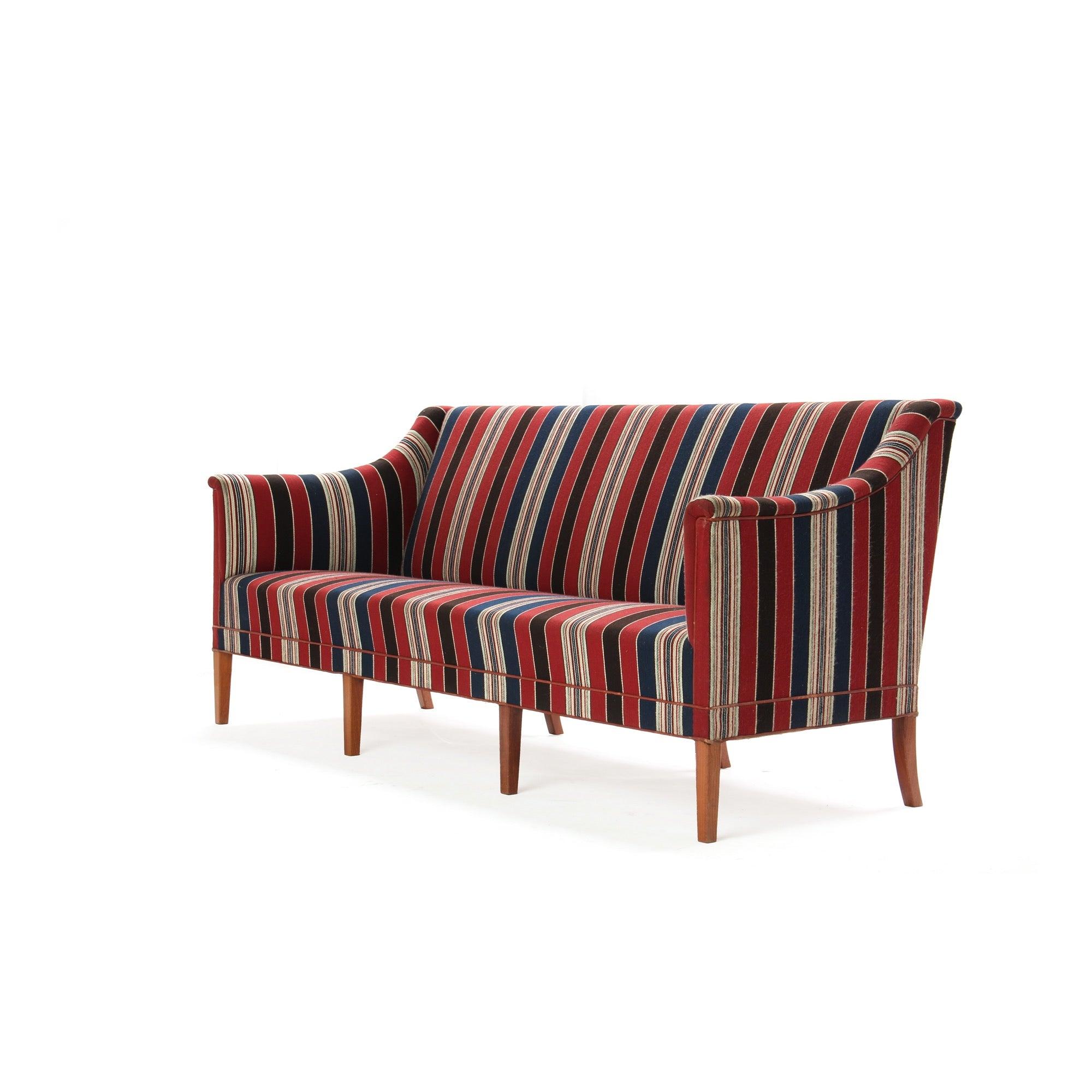 A majestic sofa having a sculptured body resting on eight (8) tapered mahogany legs, retaining the original red, navy and beige striped wool fabric. *upholstery required.