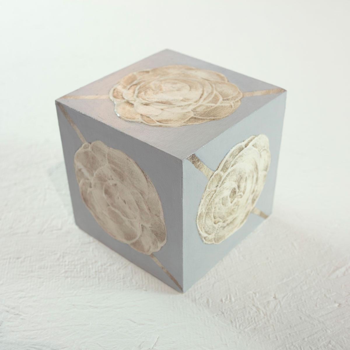 This 5" hand-painted sculptural cube by Sofie Swann is made with acrylic paint and gesso on wood. It features light texture and a warm neutral plaette, with a grey background and a large sandy beige abstracted floral form on each face of the cube.