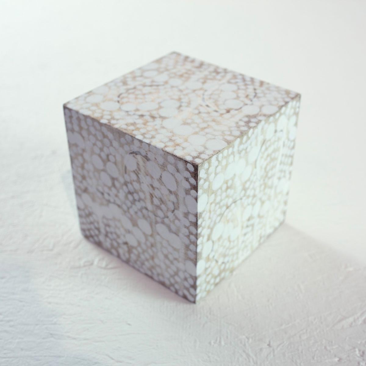 This 5" hand-painted sculptural cube by Sofie Swann is made with acrylic paint and gesso on wood. It features a sandy beige and white palette with imperfect white circular forms arranged closely throughout the surface. It is signed by the artist on