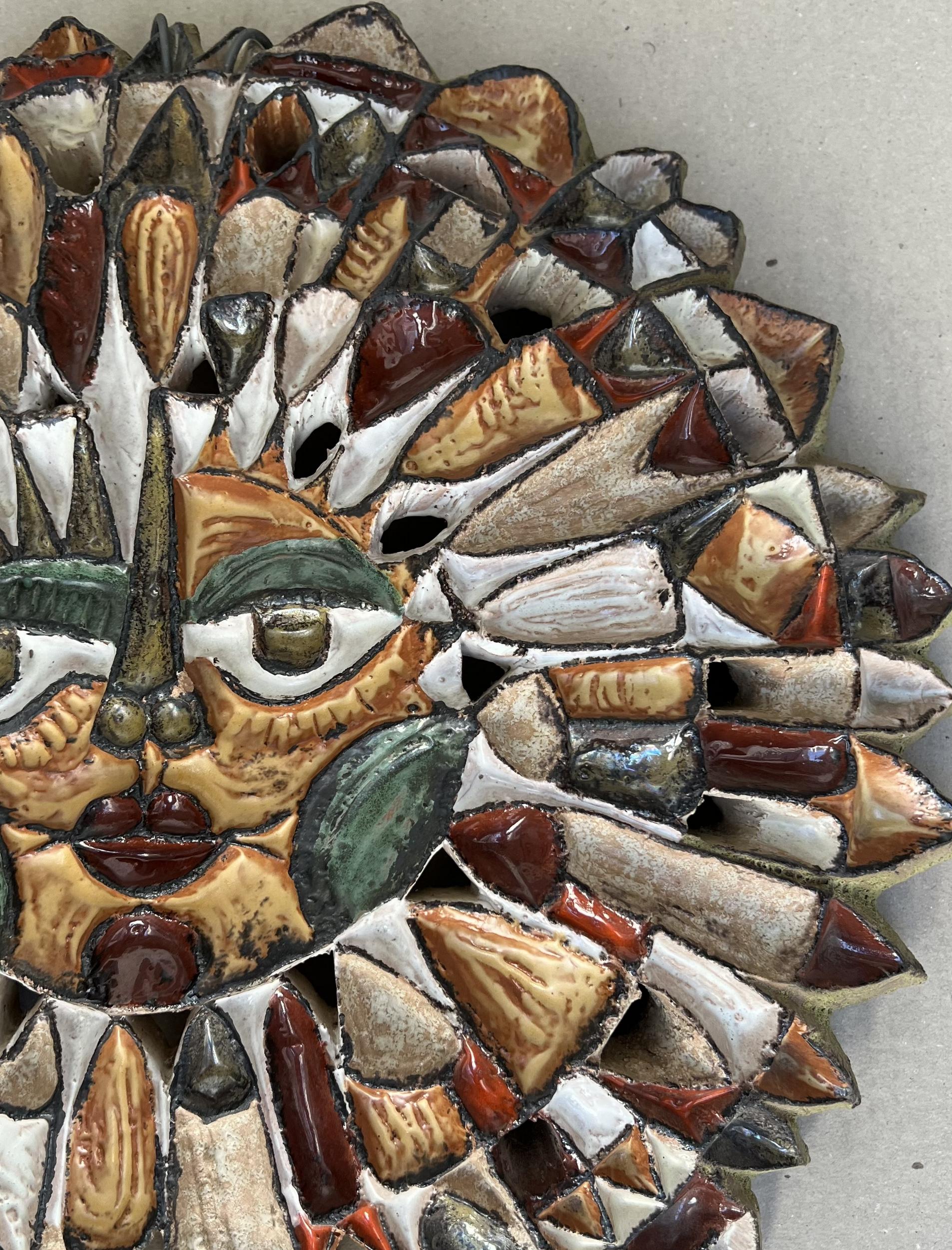 Masque solaire en céramique avec émaux polychromes. La face centrale est soulignée de jaune et de blanc. Espaces ajourés entre les différents rayons émanant du visage.

Signé 