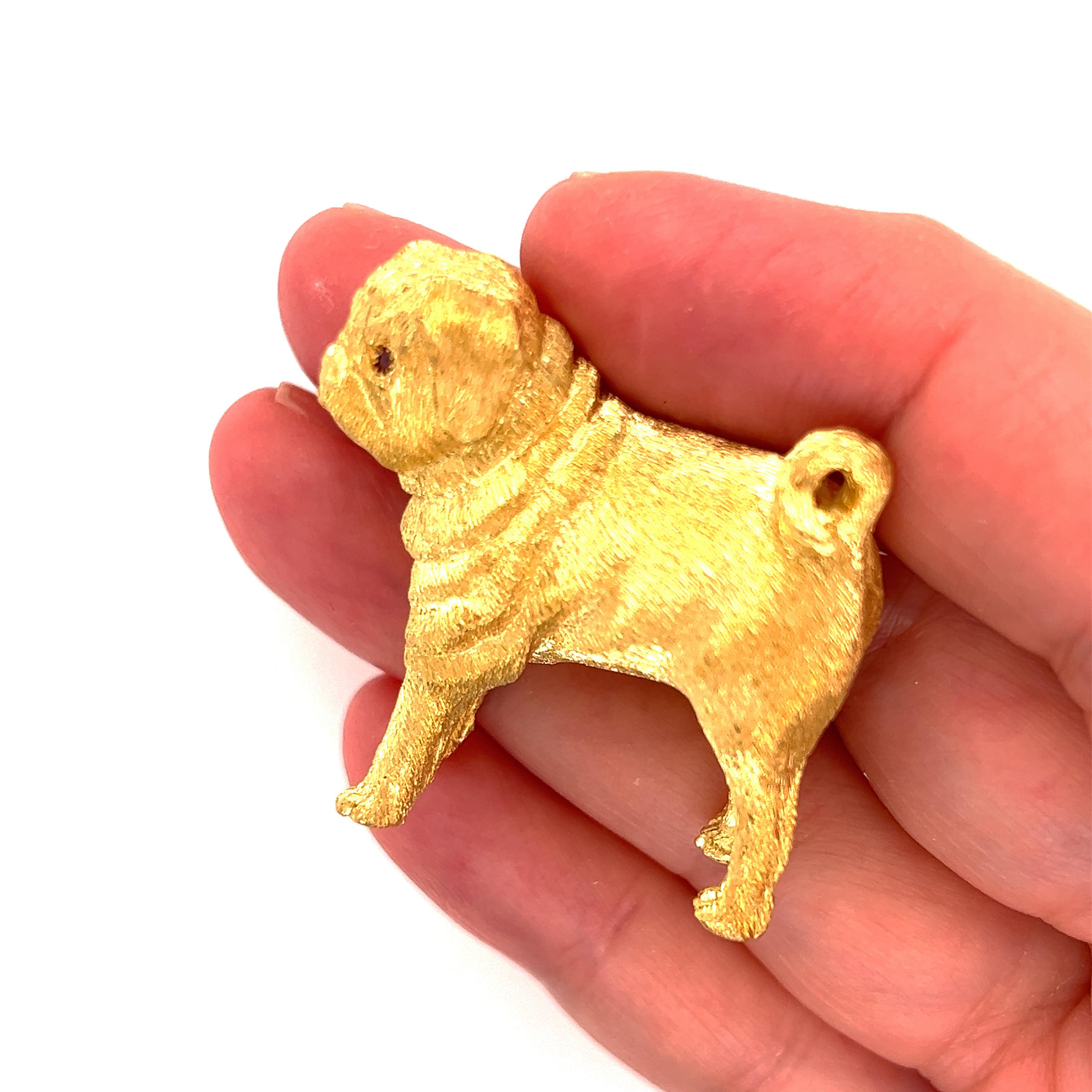 This adorable brooch is crafted from 18k yellow gold featuring a full figure standing pug dog with fur like textured finish and ruby eyes. The back of the dog comes with a safety clasp for the pin. It has the 18k gold content stamp and an arrow