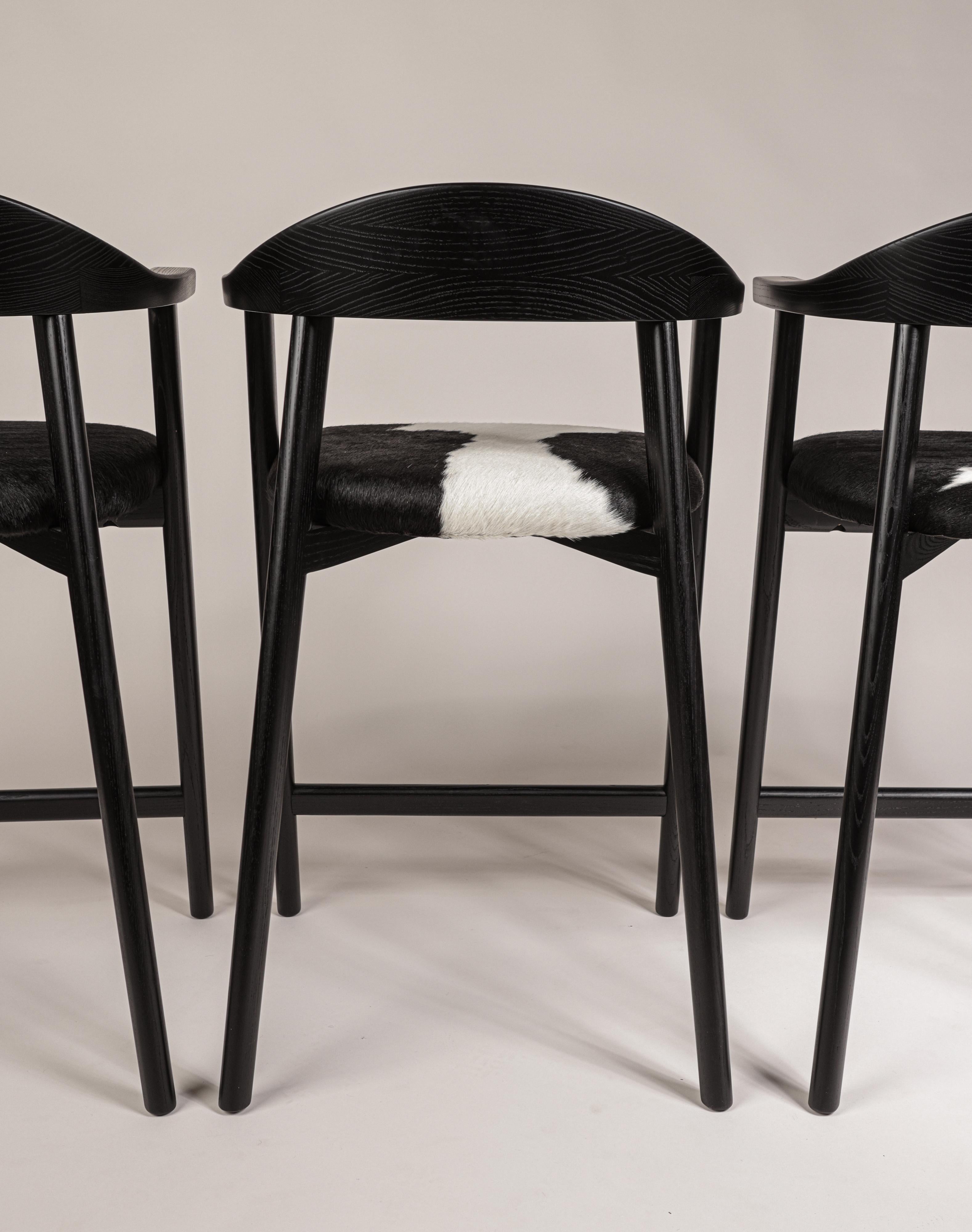 Karve Thekenhocker aus geschwärzter Esche mit schwarz-weißem Rindslederbezug. Ein klassisches Design, elegant und leicht in der Form, handgefertigt, um Generationen zu überdauern. 
Dieses Set aus vier Thekenhockern ist wie abgebildet sofort