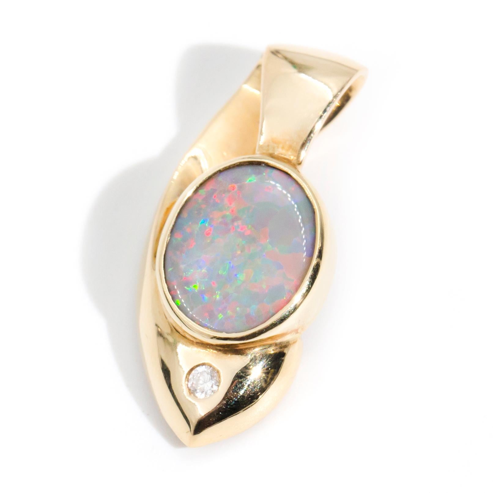 Crafted in 18 carat yellow gold, this charming vintage pendant holds an alluring opal blazen with playful splashes of red and green, resting in a classic oval setting under which lies a sparkling round brilliant diamond. We have named this luminous