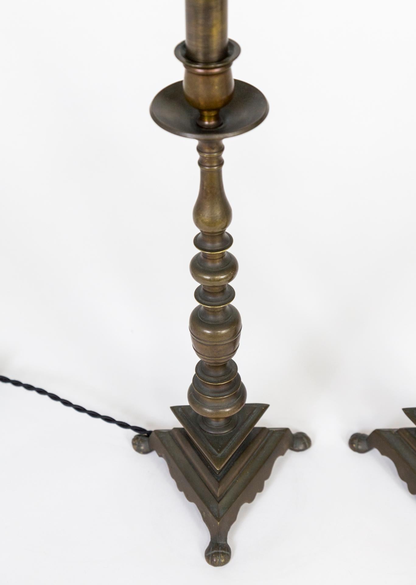 A pair of fine candlestick lamps in solid brass in a turned design with triangular bases. Candelabra sockets with matching deep bronze toned, brass candle covers, newly wired with fabric covered cord. Measures: 17” height x 6” width x 6.”
