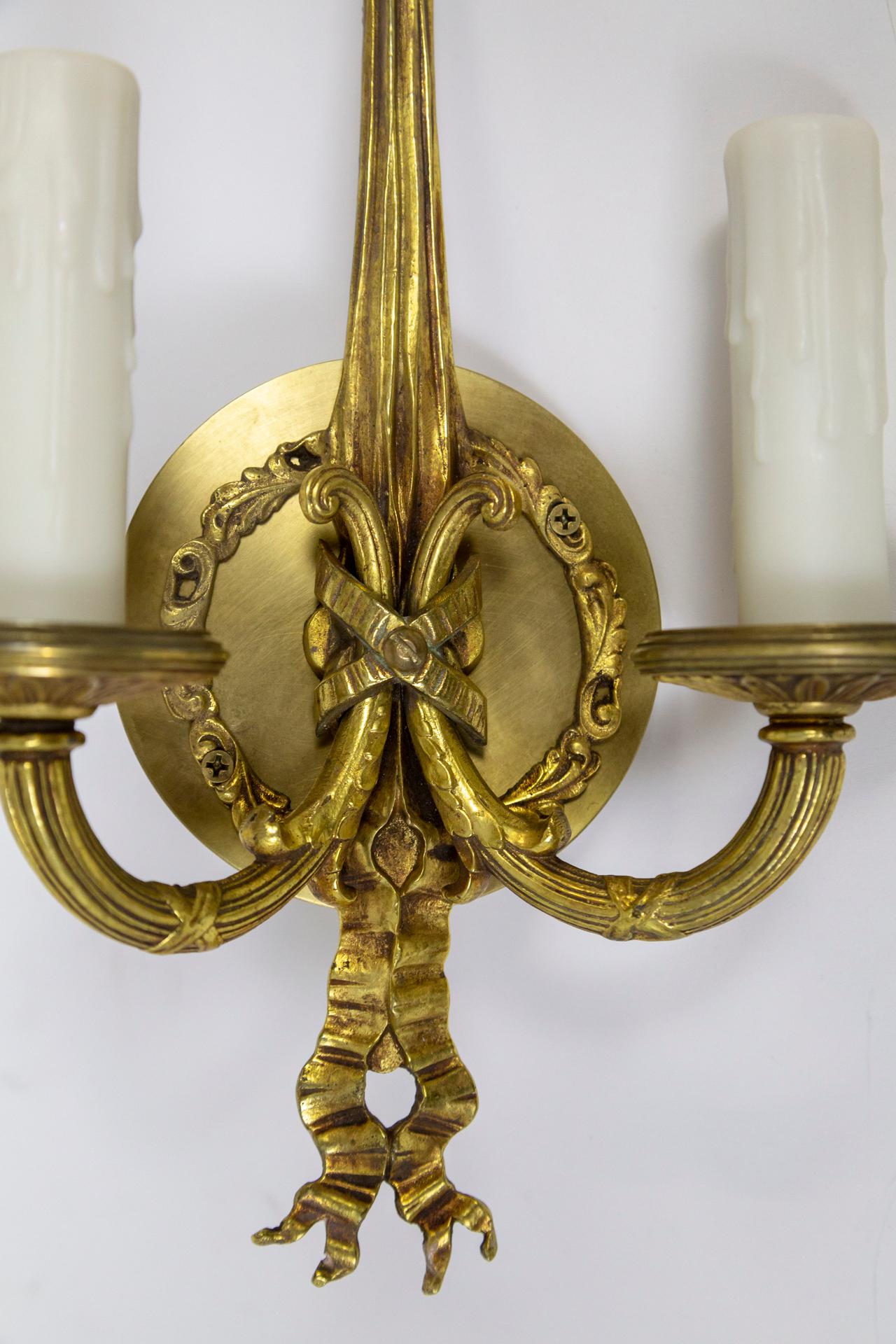 A pair of 2-arm wall sconces beautifully cast in solid brass with a reeded, pointed form, ribbons and rosettes. Made in the early 20th century in King Louis XVI style. Newly wired with faux wax (resin) candle covers. Measures: 8