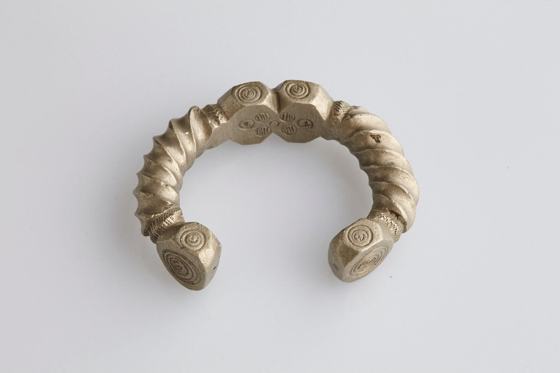 Early 20th-century brass or copper alloy with higher nickel content. Bracelet in horseshoe form with fixed opening. Turned design with rectangular shapes and carved swirling patterns. Nupe People, Nigeria.
148,00 gr / 5.25 oz. Opening 3.0 cm.
The