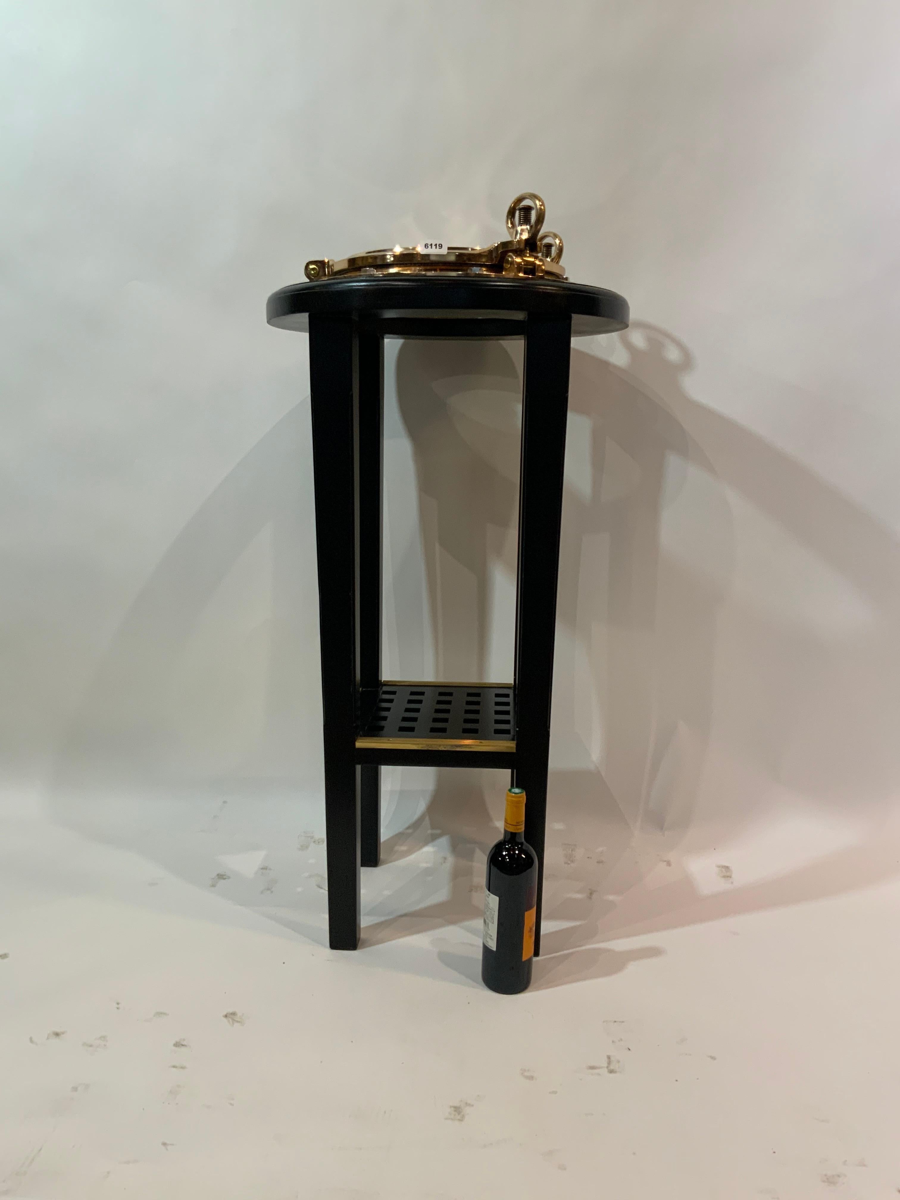 Solid brass ships porthole fitted to a bar height cocktail table stand. Highly polished and lacquered ships porthole has hinged door and two dog bolts. Stand has a ships grating shelf. Rich finish. Weight is 56 pounds. Dimensions are 40 tall by 22