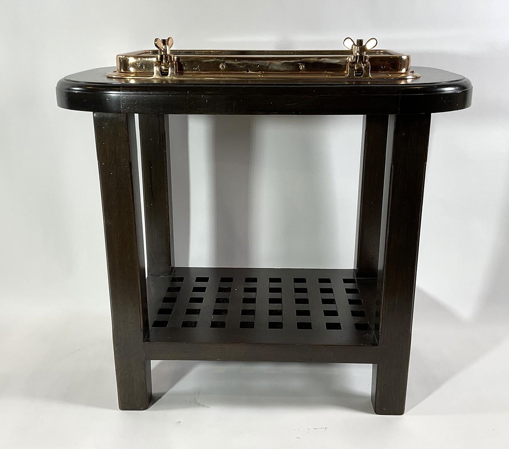 Solid brass nautical porthole table. A yacht porthole with hinged door and dog bolts has been fitted to a custom mahogany table with lower shelf in the form of a ships grating. The rectangular porthole has been polished and lacquered. Porthole is