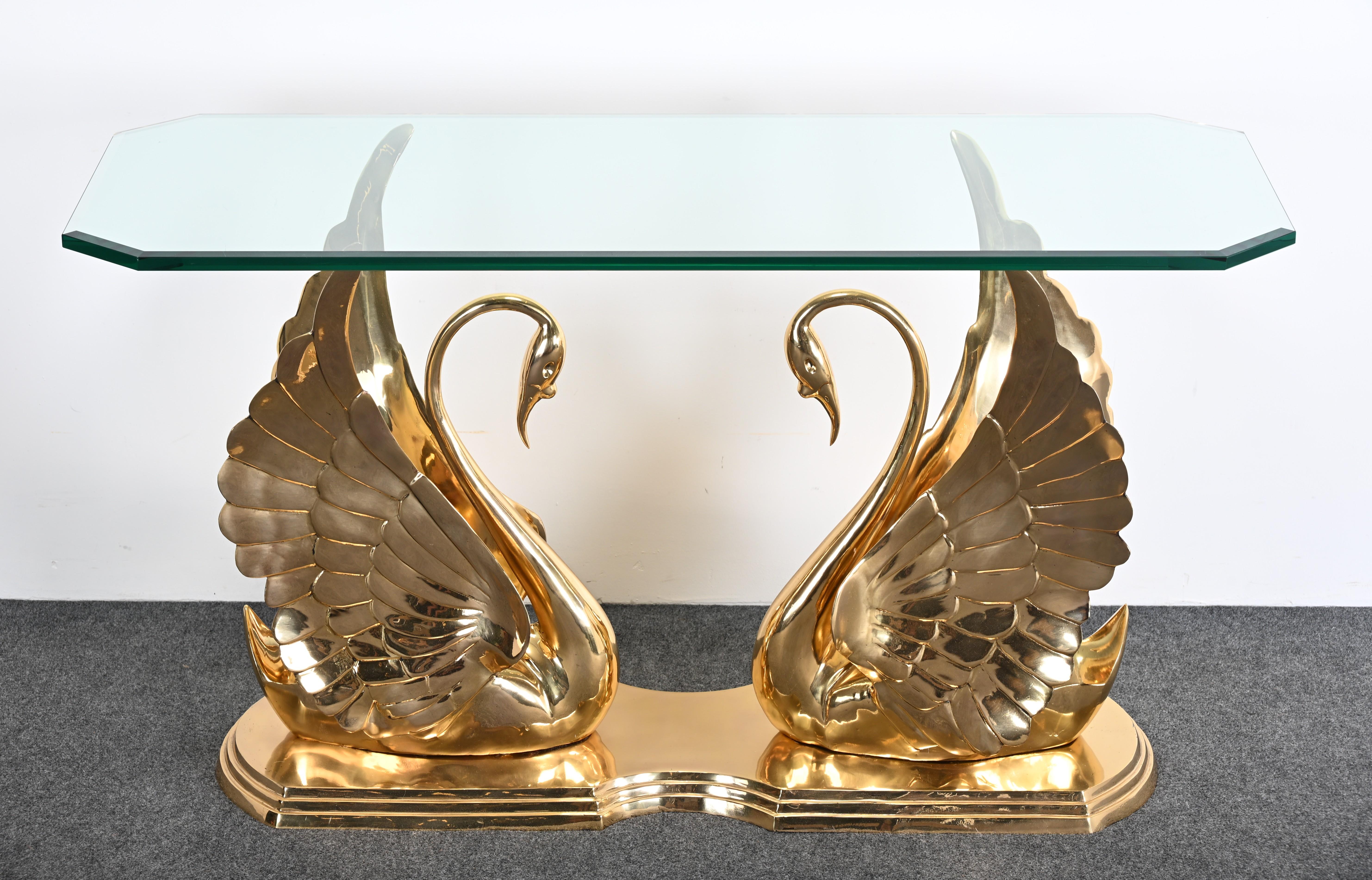 The image shows a solid brass swan console table with a glass top, which appears to be designed in the manner of Maitland Smith. This style is characterized by its ornate, handcrafted details and luxurious materials, often featuring brass and