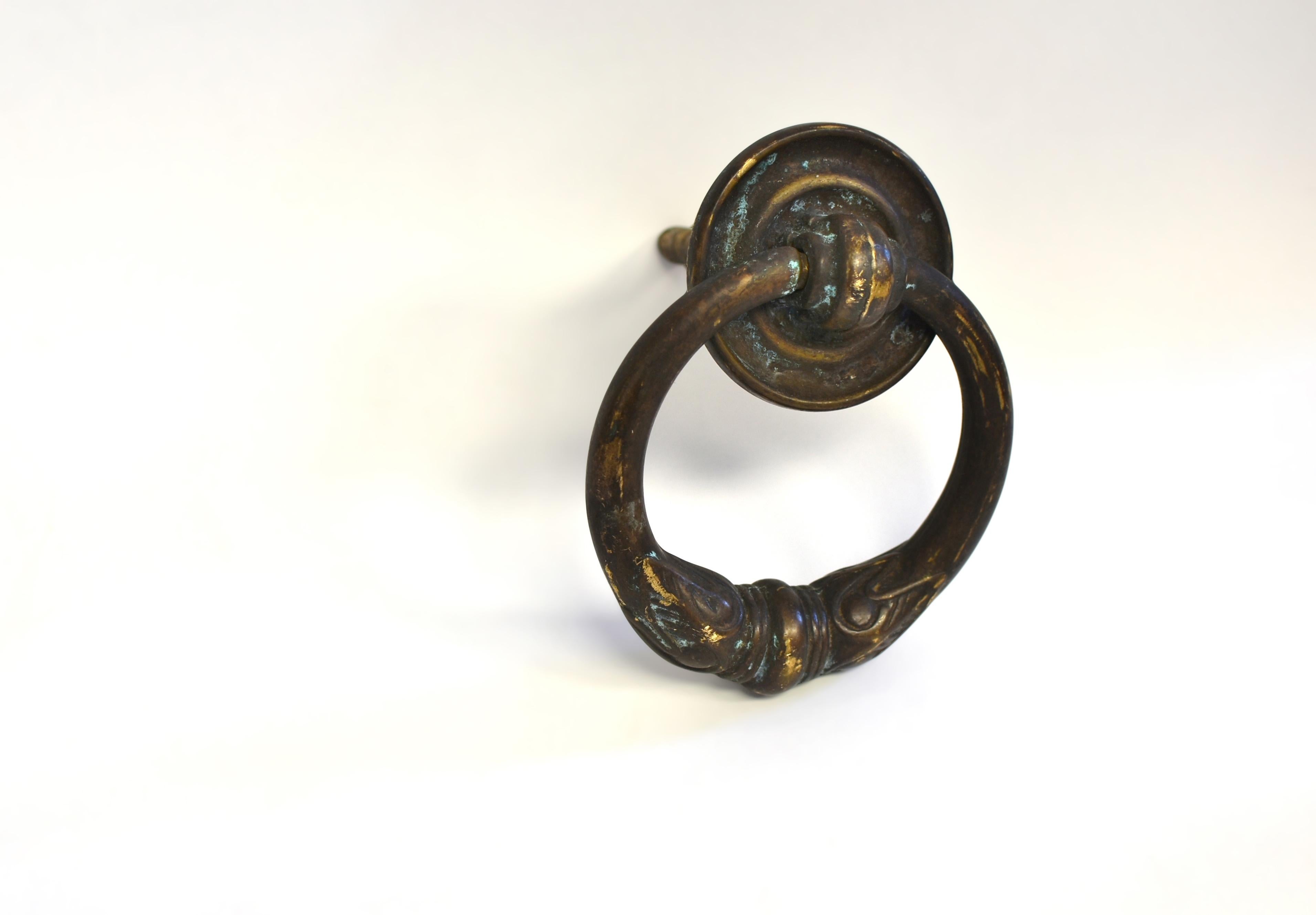 A solid antique bronze door knocker in the form of a circular handle, gradually widen reaching the middle. A pair of heraldic emblems depicting eyes with feathers decorate the knocker at the center bottom. The substantial knocker is surmounted by a