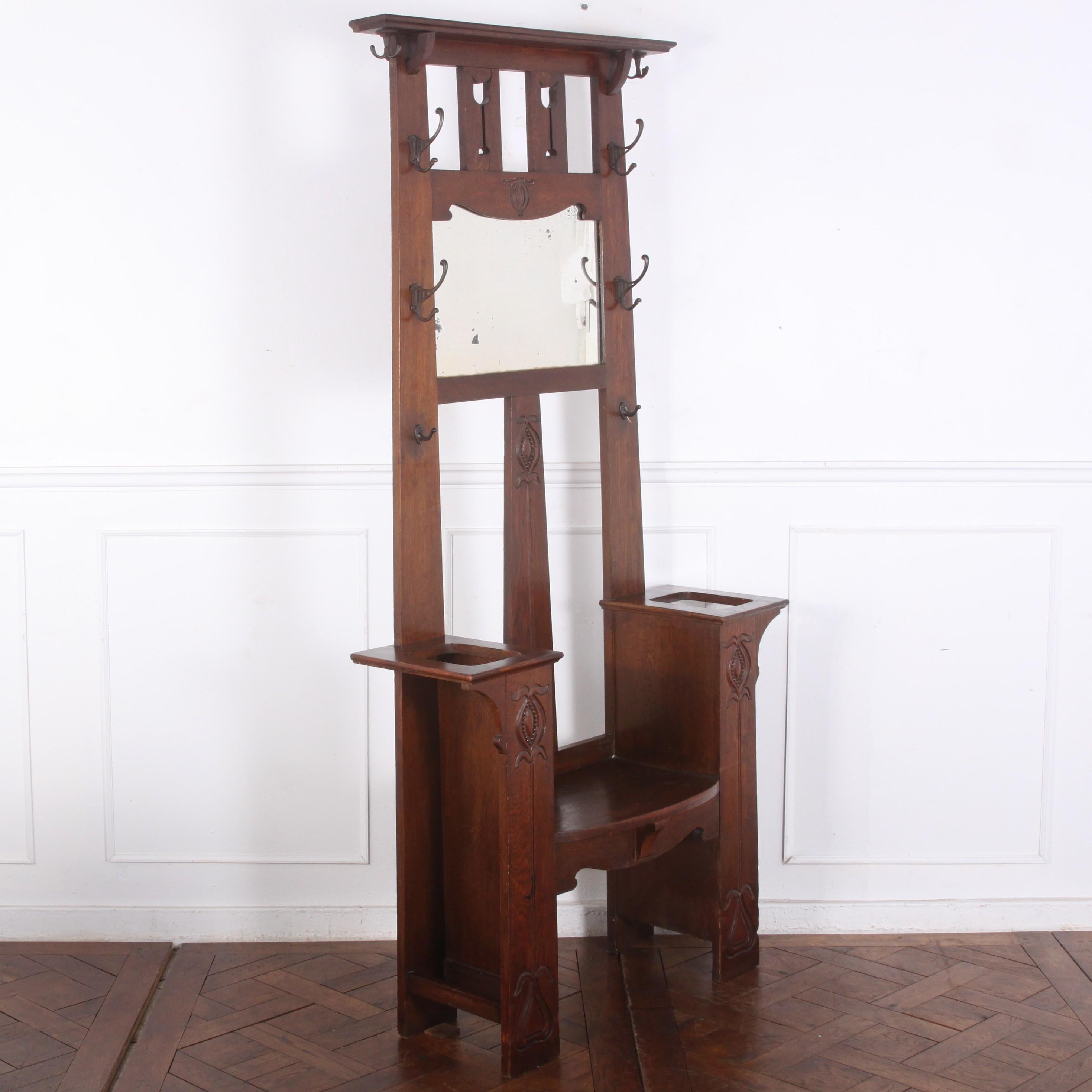 A British- possibly Scottish- carved oak Arts & Crafts period seated hall stand with simple geometric lines typical of the style and abstracted ‘pomegranate’ carved details. Upper mirror with a variety of hooks and side slots for walking sticks and