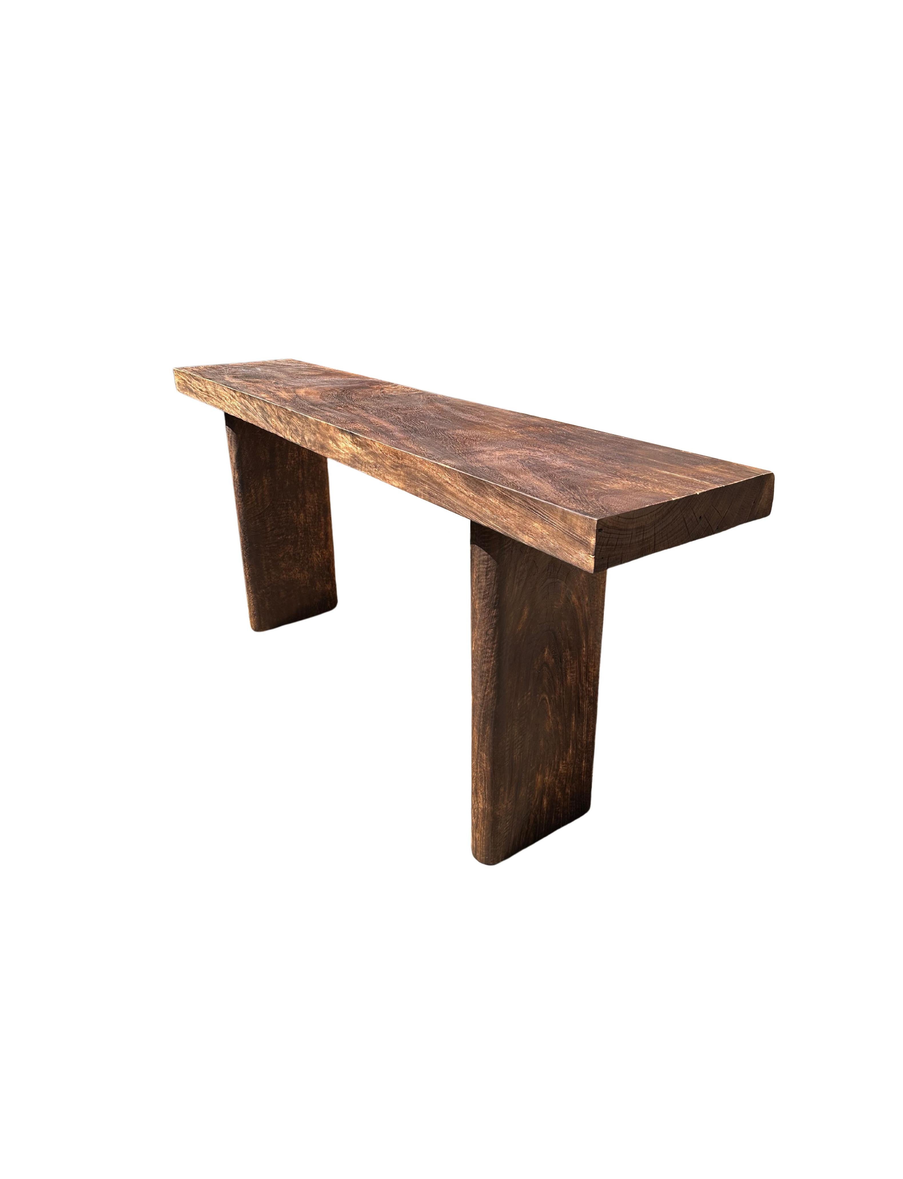 A sculptural mango wood console table crafted from a solid block of mango wood. The table top sits atop two solid, narrow legs. The tables legs feature curved edges providing a wonderful mix of curved and straight lines to the table. A wonderfully