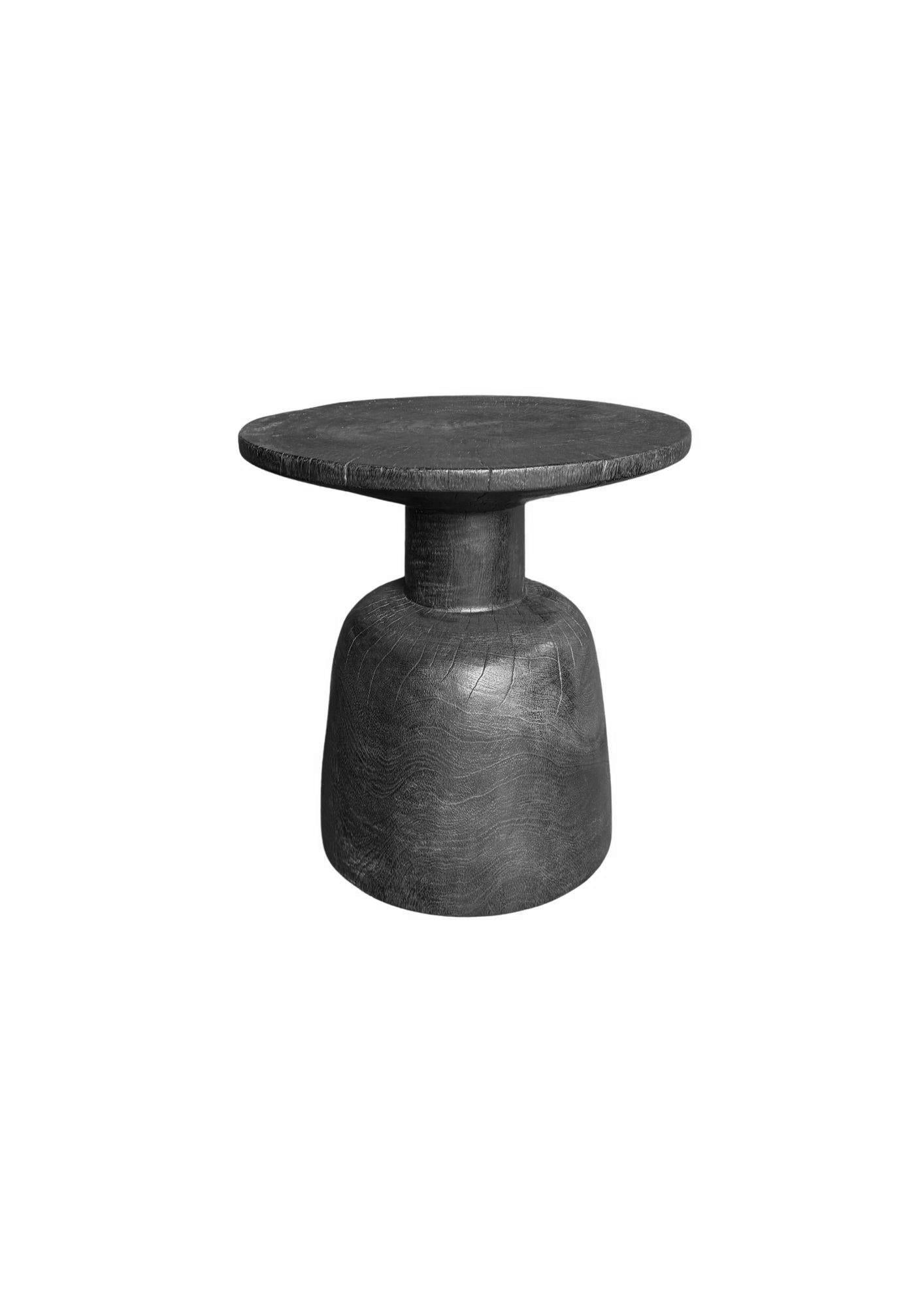 Cette table d'appoint remarquable est fabriquée en bois massif et a été brûlée plusieurs fois de manière experte pour obtenir un pigment noir riche et profond. Ce procédé met en valeur la texture naturelle du bois, la surface présentant des motifs