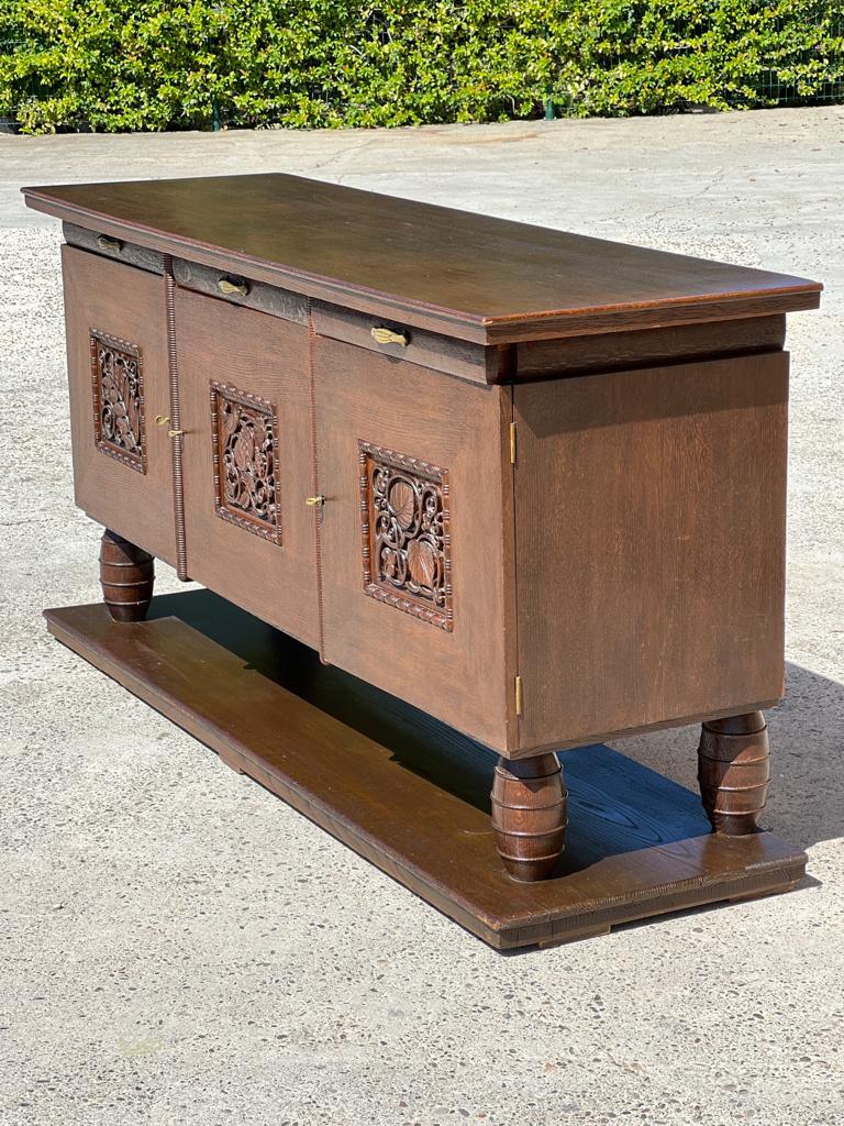 Buffet en chêne massif teinté dans le style de Joseph Savina 1940. 
Ouvrant sur 3 portes sculptées d'oiseaux et de fleurs, et 3 tiroirs avec poignées en laiton. 
Plateau rectangulaire légèrement débordant et pieds d'angle en forme d'olive sur une