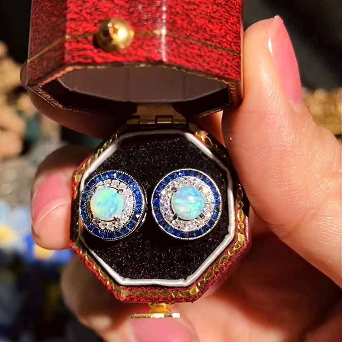 Questi orecchini Art-Deco sono assolutamente spettacolari! I diamanti scintillanti e i vibranti zaffiri blu circondano l'eccellente gioco di colori dell'opale centrale, che si trova in un sottile castone con dettagli millgrain. Realizzato in oro
