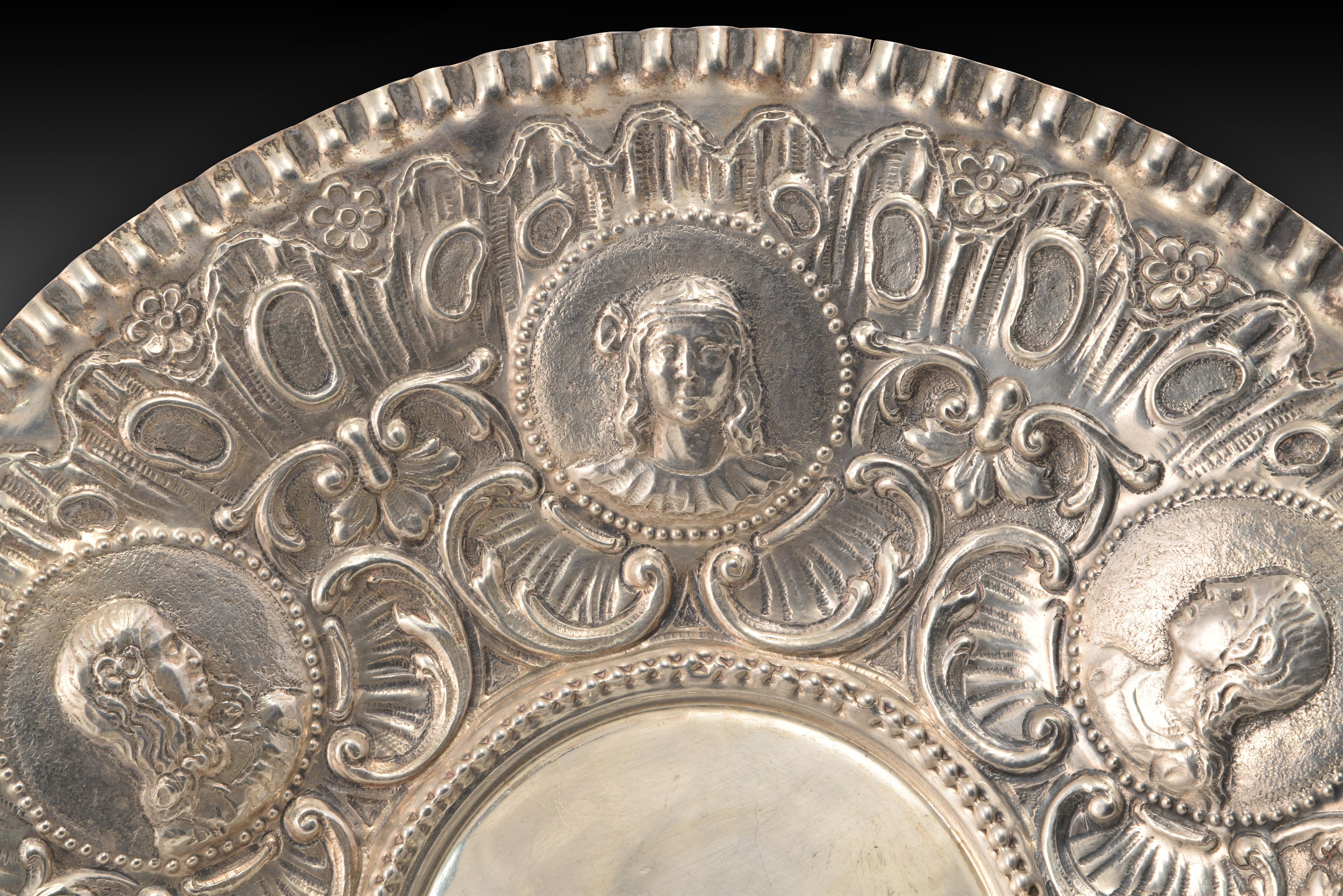 Circular silver tray embossed in its color, circular and decorated on the edge with a series of busts inside pearl strings, accompanied by scrolls, plant elements and motifs of clear Rococo memory, which has a jagged edge to the outside. The piece