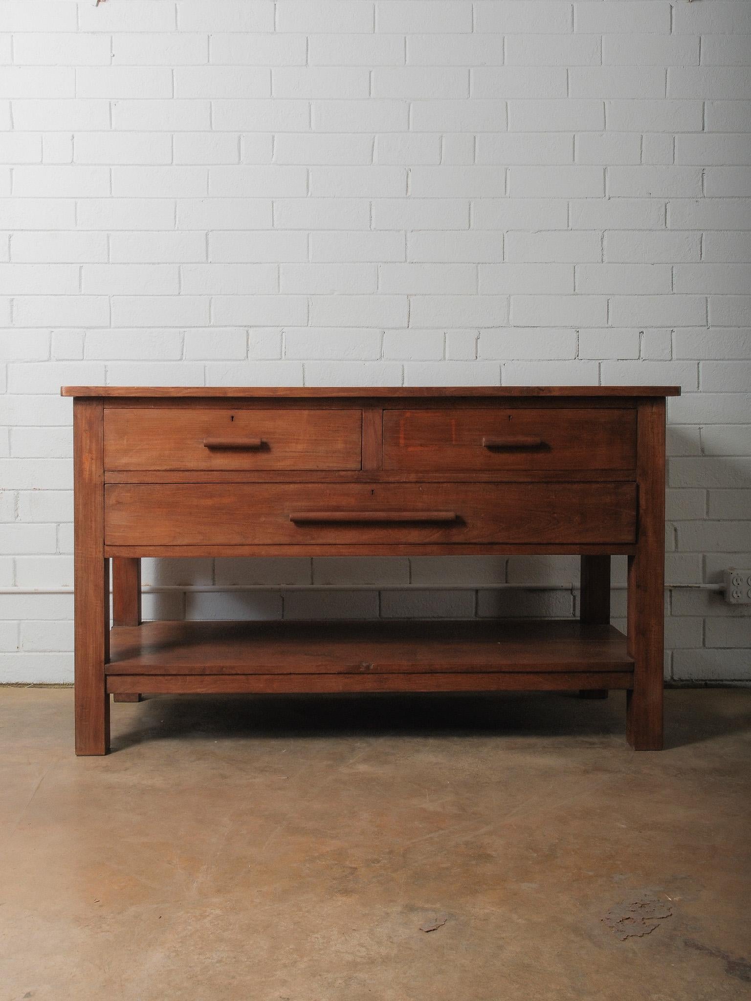 A substantial solid wood work table defined by its clean lines, honest joinery, and warm, matte patina. The surface reveals soft tonal variations in the grain—deep honey, umber, and understated red-brown—creating a natural depth that feels both