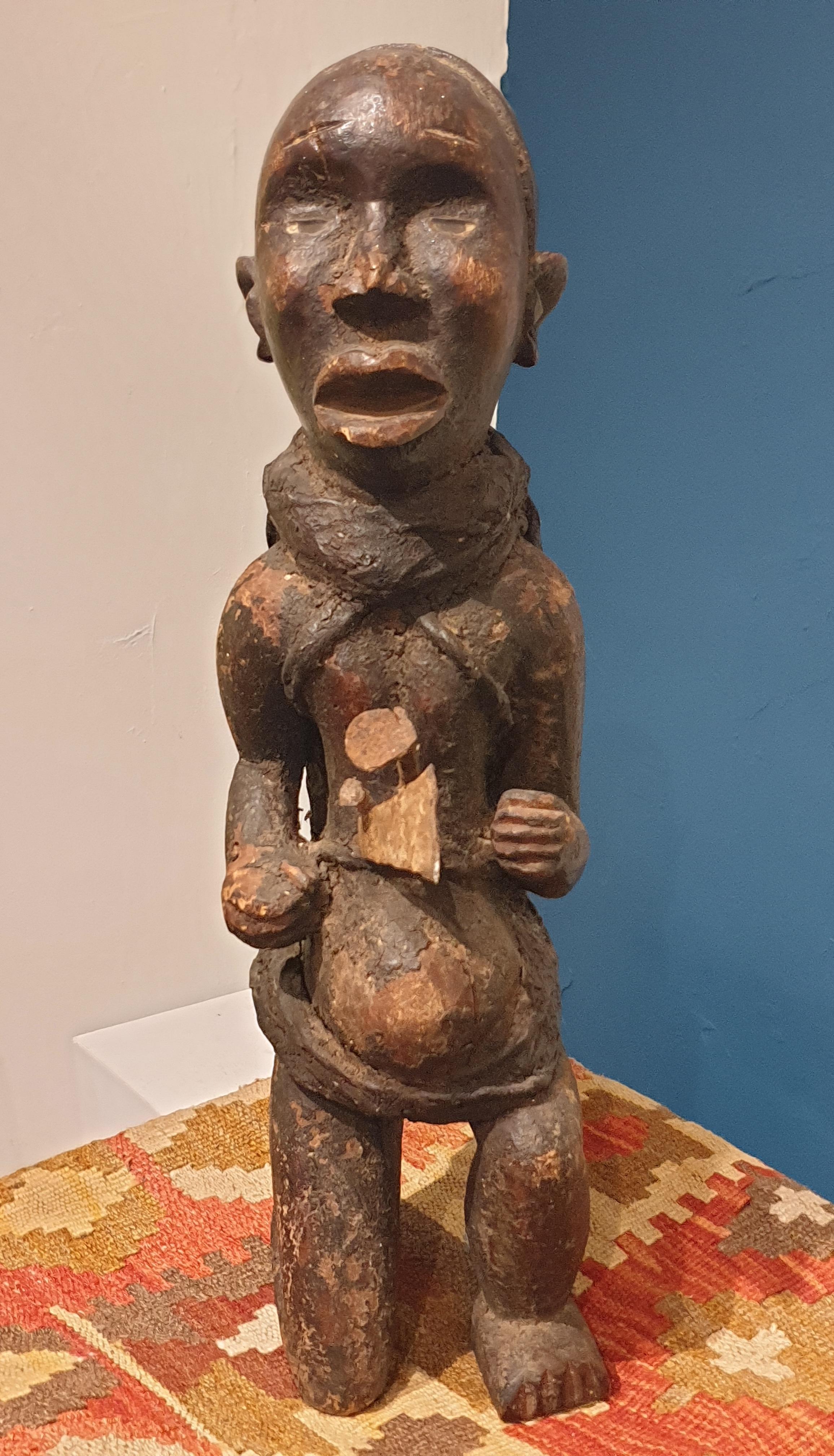 A Songye Male Power Figure, Democratic Republic of the Congo, the male figure resting on one knee, with openwork arms and hands resting by the abdomen which contains various charms and hand forged items, carrying on his back a bound 'hote'. Typical