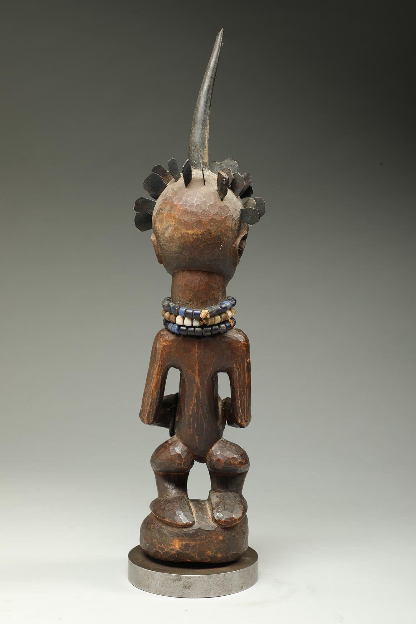 Carved wood standing female figure with projecting stomach, attached horn on top of head, and hair in form of small hand shaped copper pieces. Old glass beads around arm and neck.
It is in good condition wear and erosion from traditional use,