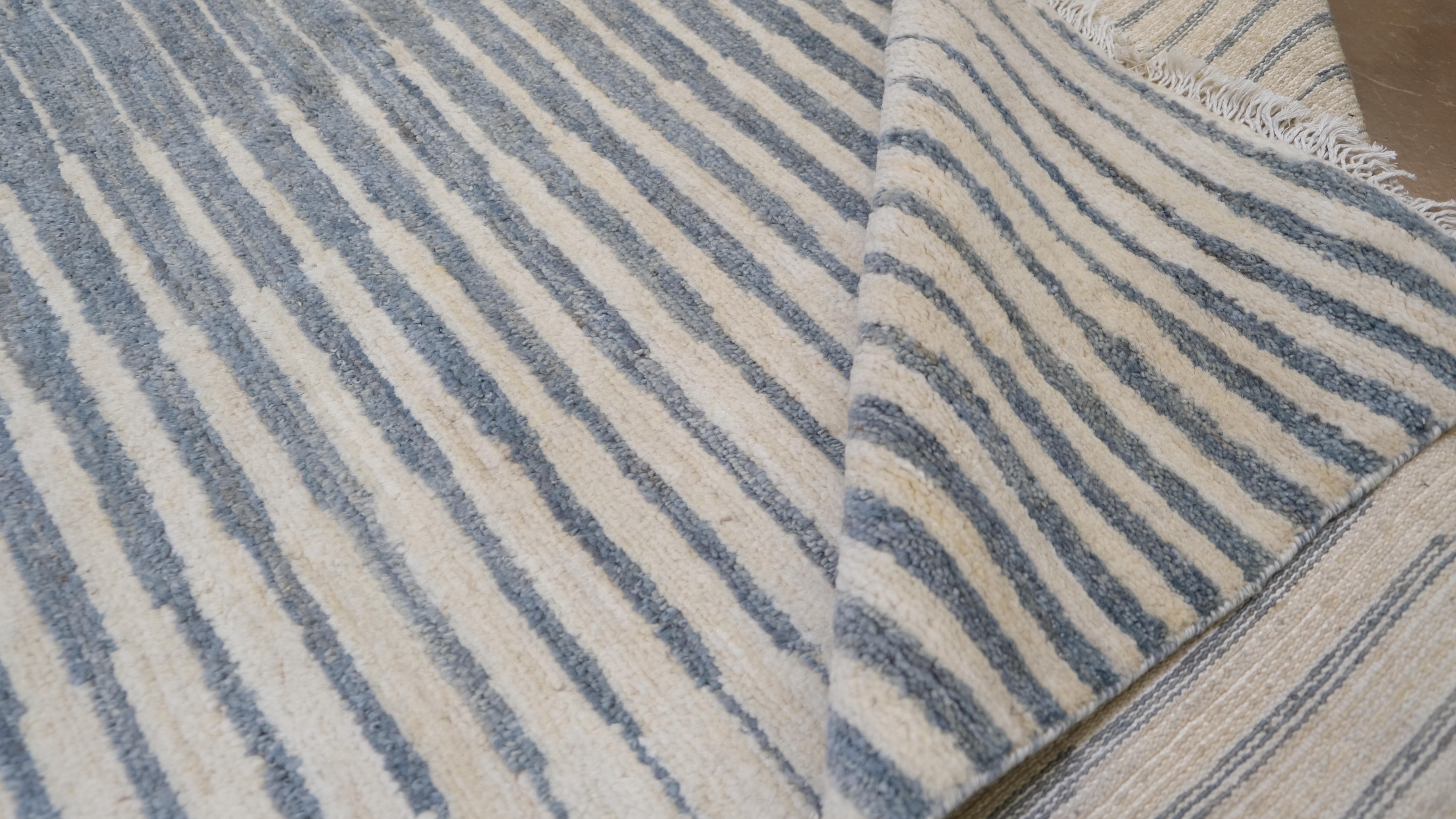 Bringen Sie subtile Eleganz in Ihr Zuhause mit diesem atemberaubenden handgeknüpften Wollteppich. Mit seinem beruhigenden Streifenmuster in Blau-, Beige- und Elfenbeintönen bietet dieser Teppich eine klare, moderne Ästhetik. Sein rhythmisches,