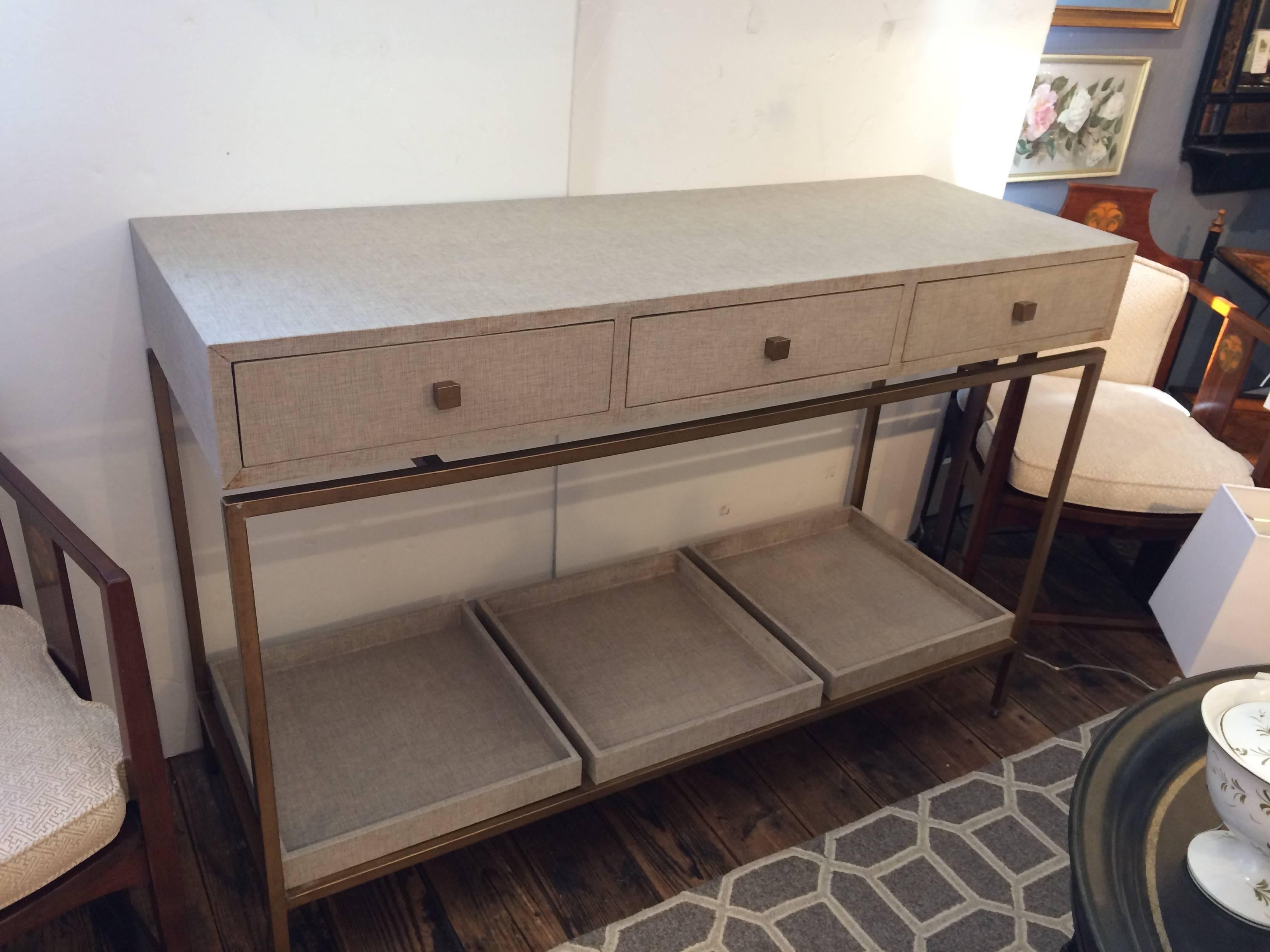 A great looking contemporary console covered in a neutral greyish beige linen (or fabric like linen) offset stylishly with gold metal base and knobs on the three drawers. A row of three removable trays make up the bottom tier.