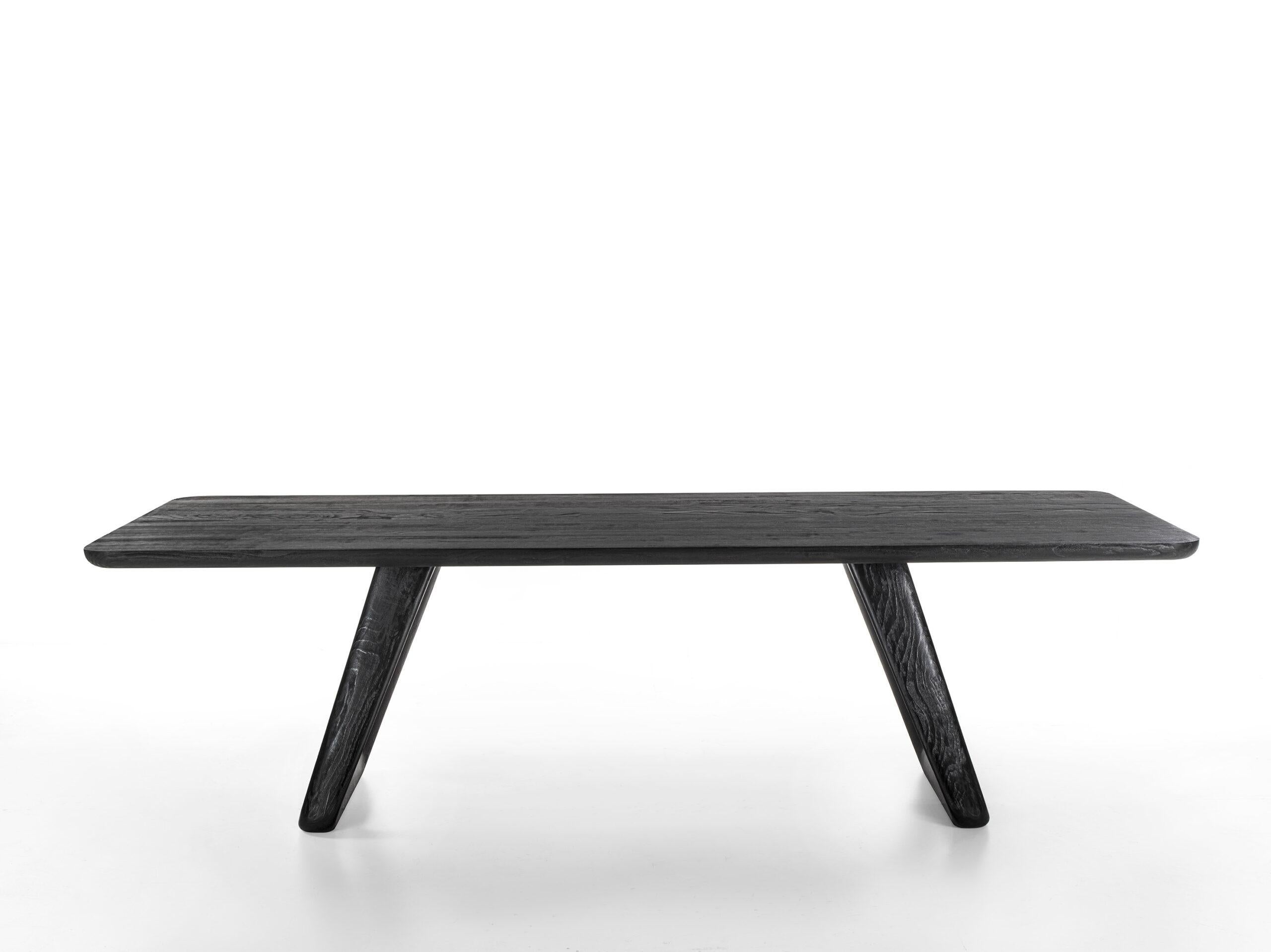 Tisch mit verleimter Massivholzplatte und abgeschrägten Kanten, charakterisiert durch zwei Beine aus Massivholz, deren raffinierte Geometrie durch das Zusammentreffen von geraden Linien mit geschwungenen Linien und abgerundeten Ecken bestimmt