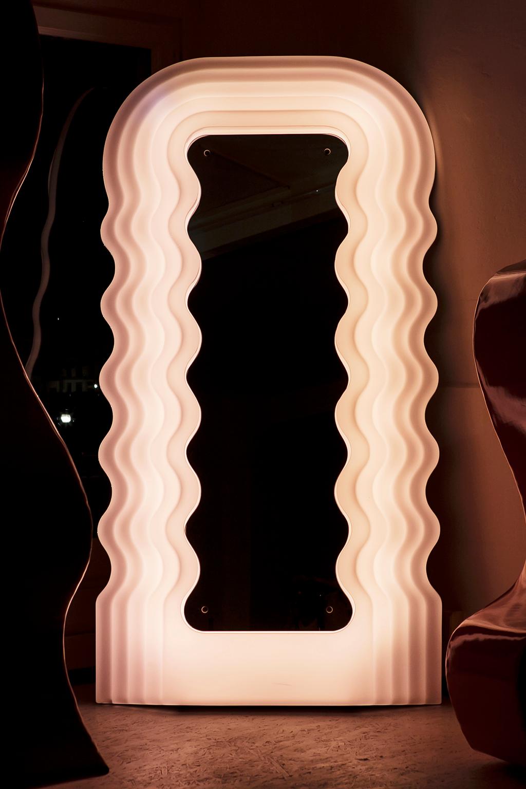 El espejo-lámpara Ultrafragola, con su perfil sinuoso que sugiere una larga cabellera ondulada, pertenece a la serie Mobili Grigi de mobiliario completo de dormitorio y salón diseñada por Ettore Sottsass para Poltronova y presentada en la tercera