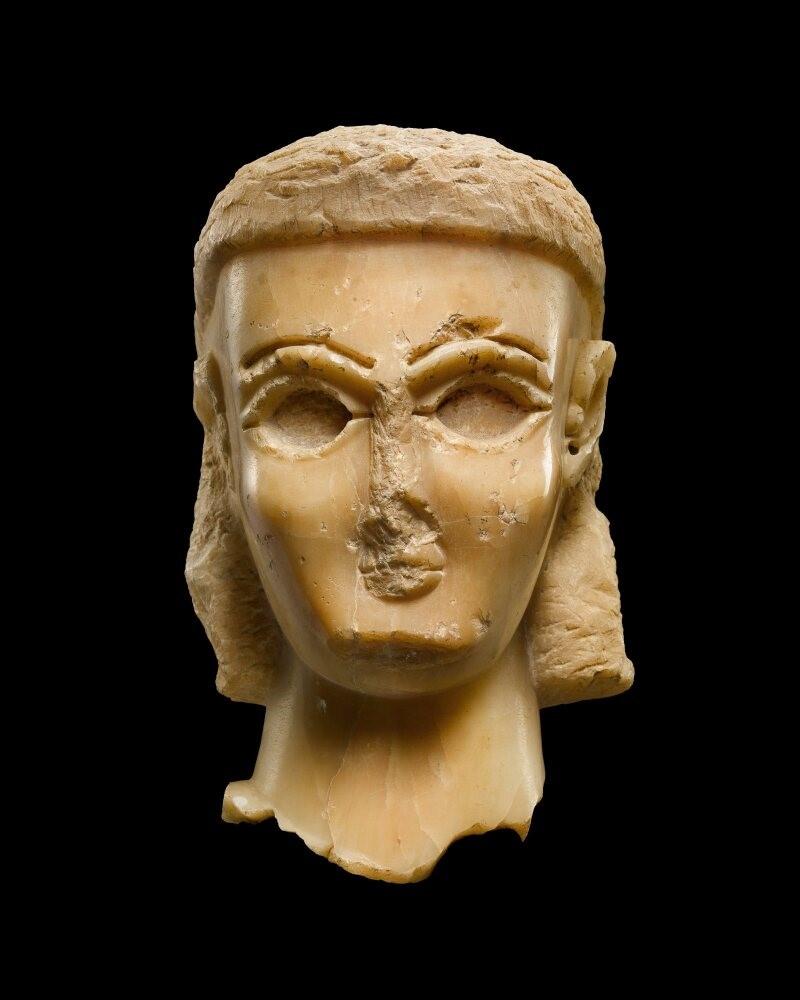 A female head finely carved from evenly-coloured cream alabaster. The face features a strong chin, high rounded cheekbones, and large elliptical eyes deeply inset for inlays. The remains of the nose indicate that it was long, thin, and roughly