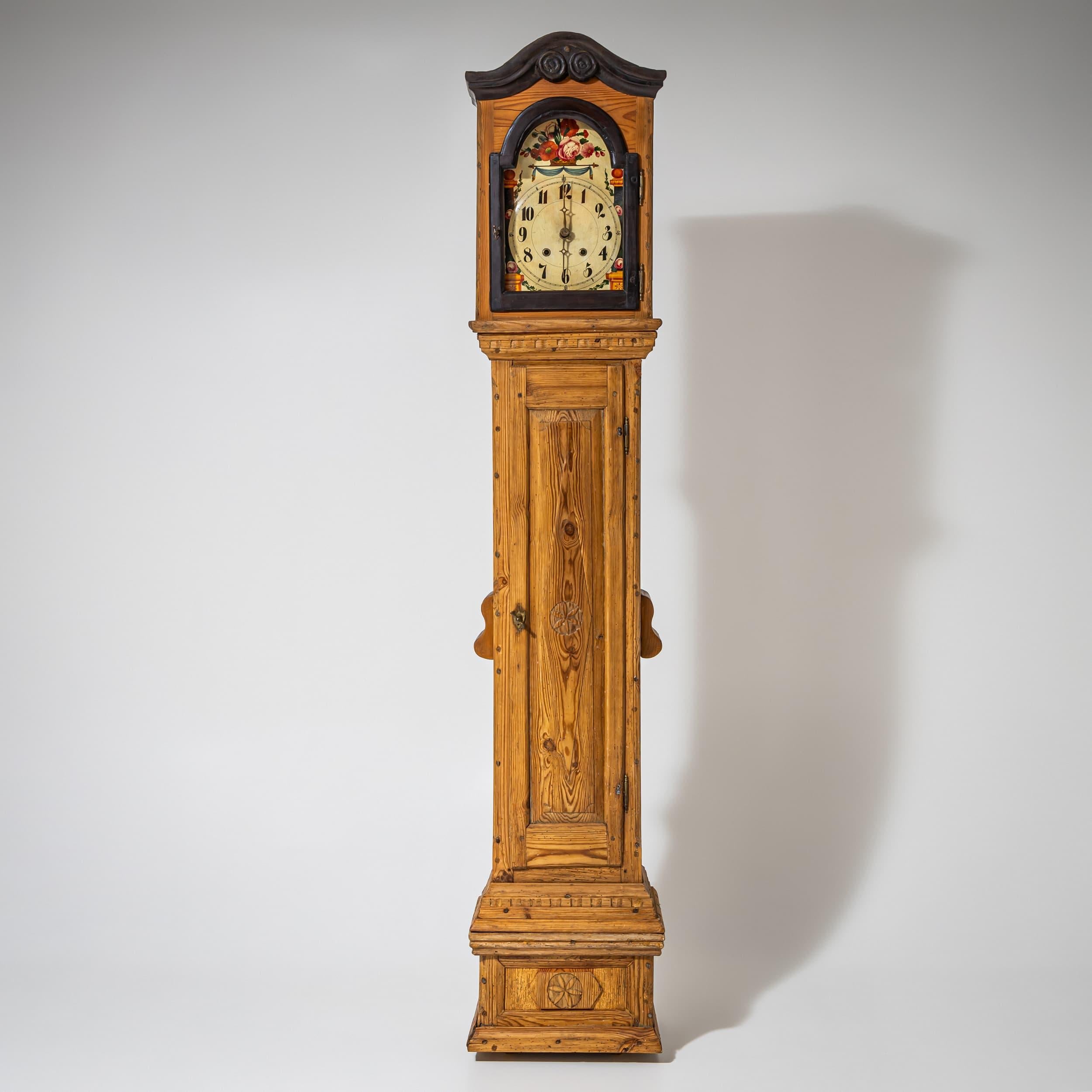 Slim longcase clock in fir wood with profiled base and tall rectangular case. The hood is framed by a curved pediment and displays a dial with hand-painted numerals and floral decoration. Side openings provide ventilation for the movement, while the