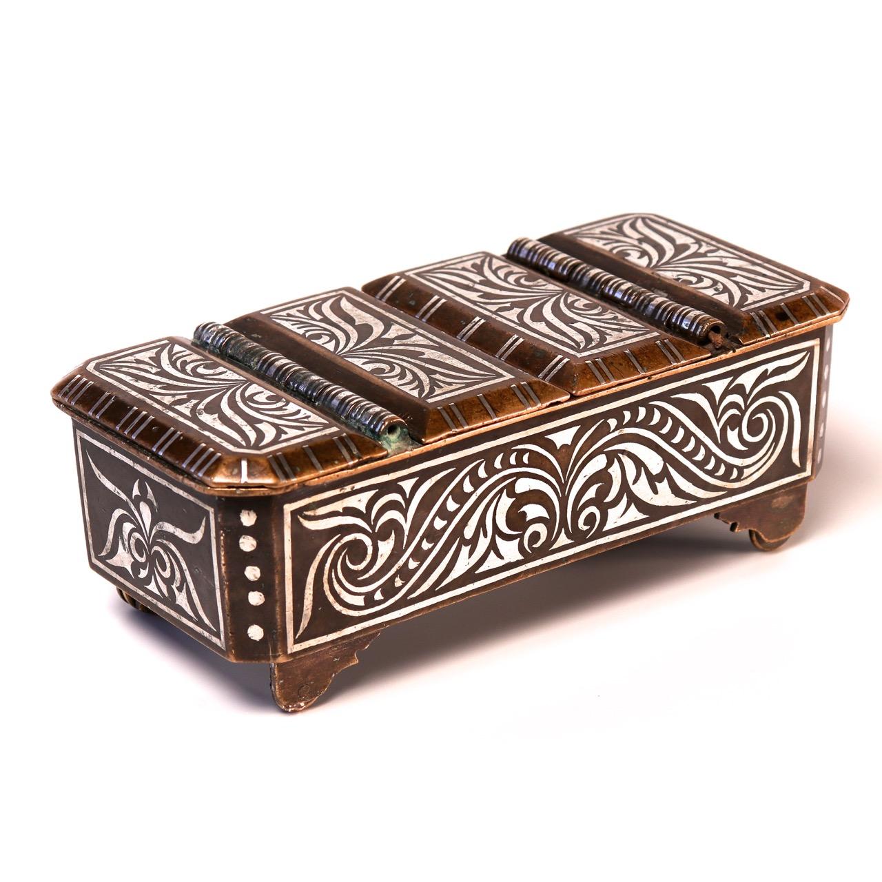 Southern Philippine (Mindanao) brass with silver inlay betel box, a compartmentalised rectangular body with four hinged lids covering three internal compartments, supported on four wheeled feet, the silver inlay on the sides, chamfered corners and