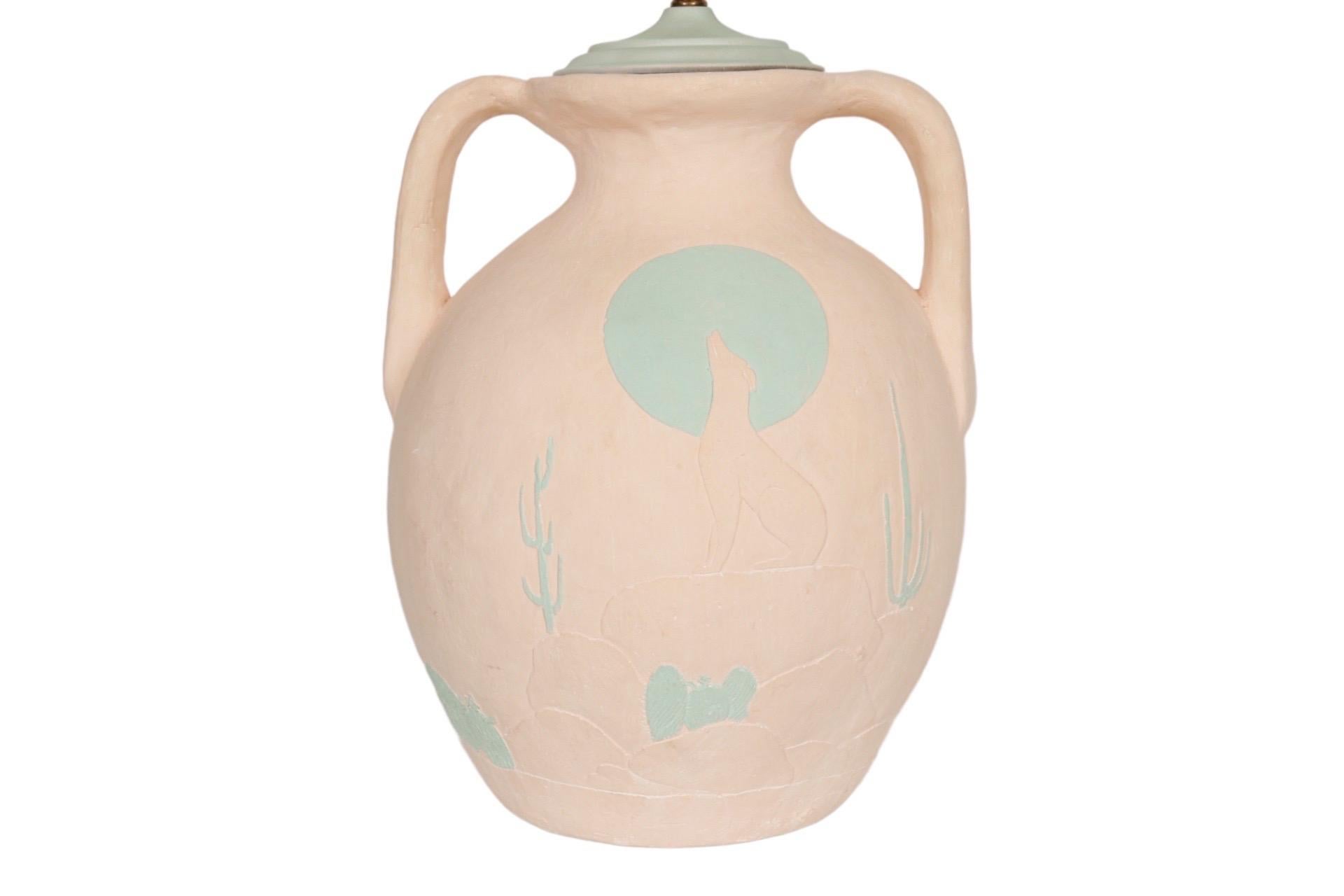 A southwestern earthenware amphora table lamp. Hand crafted, with a pink finish and decorated on both sides with an embossed and engraved desert scene. A coyote howls in front of a sea green moon, surrounded by pink sand dunes and green cacti. The