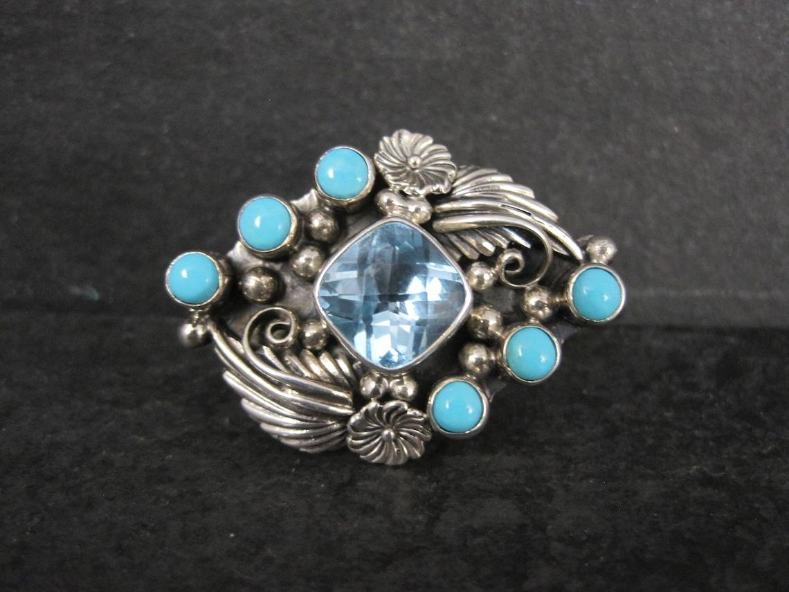 This gorgeous ring by Running Bear is a two finger ring.
It is sterling silver with a 10mm blue topaz and 6 snake eye cut turquoise gemstones.

The face of this ring measures 1 1/16 inches north to south and 1 7/16 inches east to west.
Sizes 9 and 8