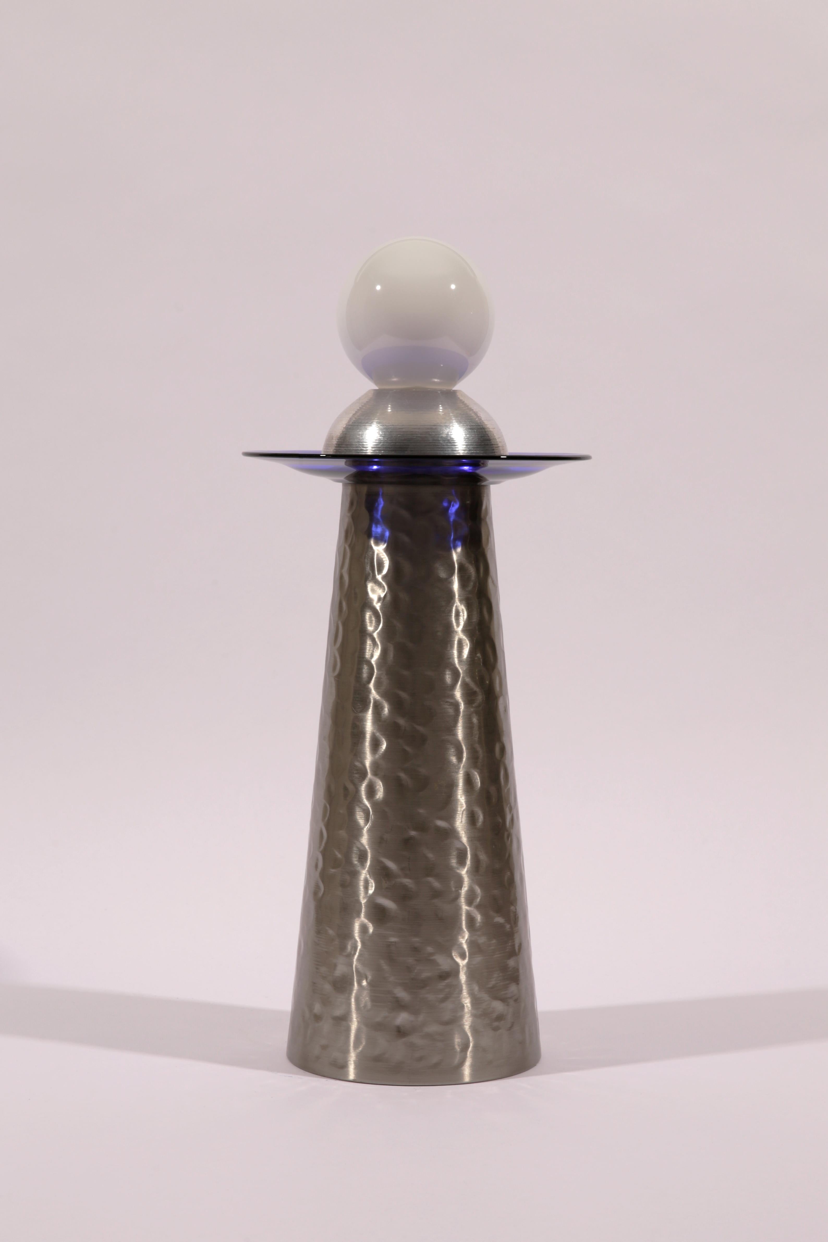 The Shape of Things to Come 2 est une lampe de table ou de sol inspirée par l'ère spatiale. Son apparence élégante et futuriste est obtenue grâce à l'utilisation d'aluminium et de verre bleu, ainsi que d'une grande ampoule à gradation non couverte.