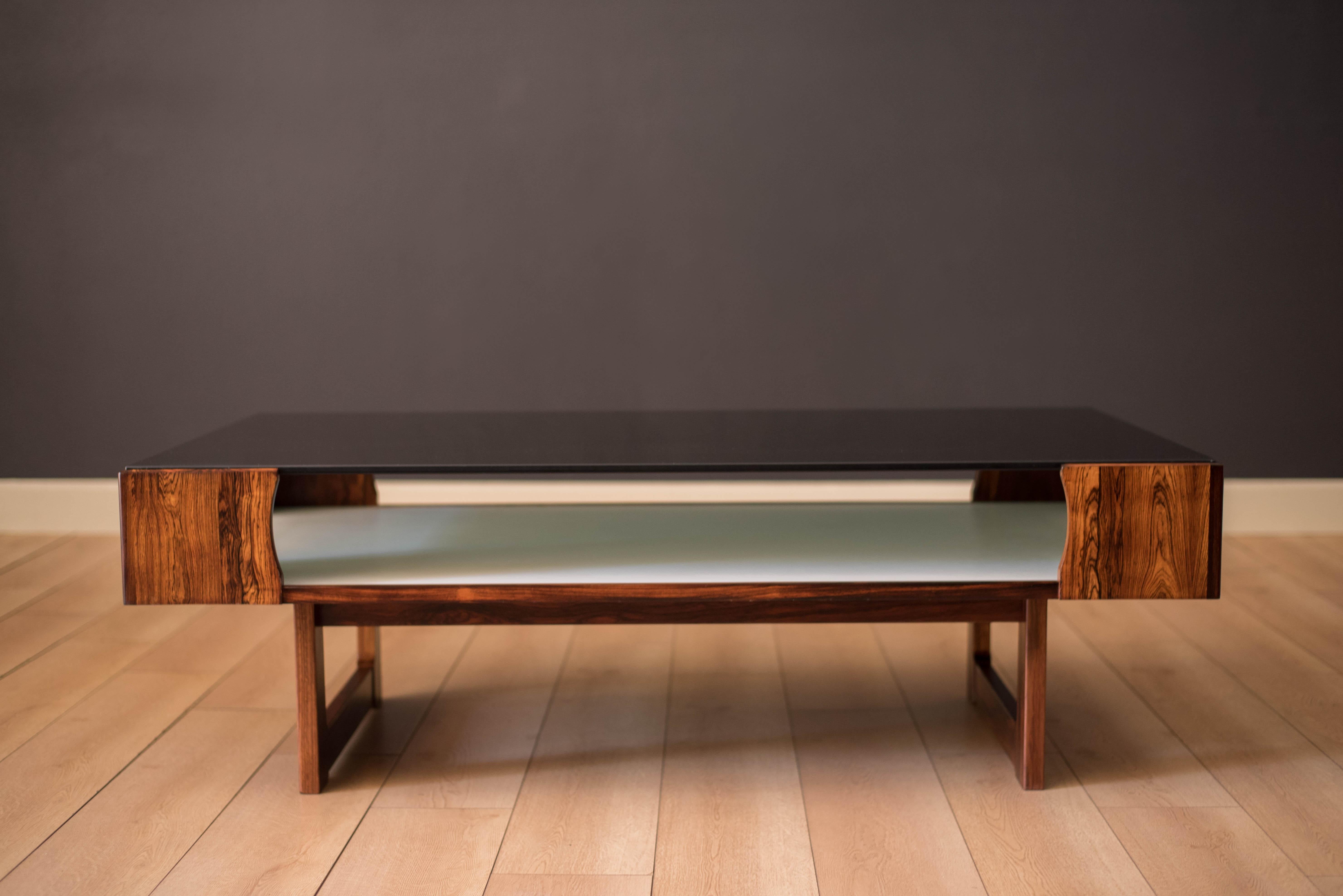 Vintage two tier coffee table in rosewood designed by Torbjorn Afdal for Bruksbo, Norway c. 1970's. This piece showcases from all angles featuring a smoked glass table top with a lower magazine shelf in white laminate for extra storage and easy