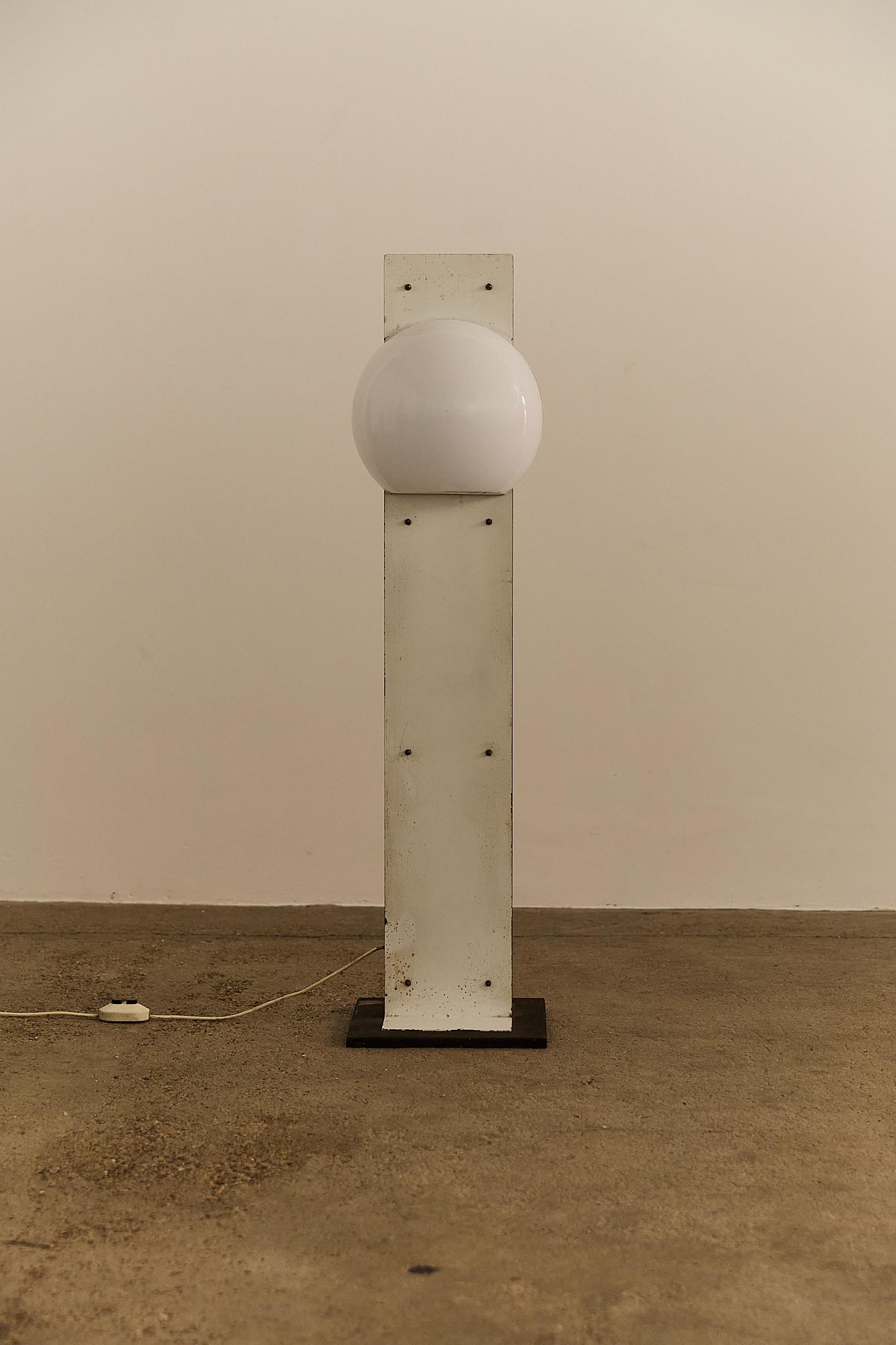 The piece blends industrial materials with futuristic form: a vertical metal support, showing visible rust and heavy patina, frames a bisected white acrylic globe. The square base and exposed bolts enhance its raw, utilitarian character, while the