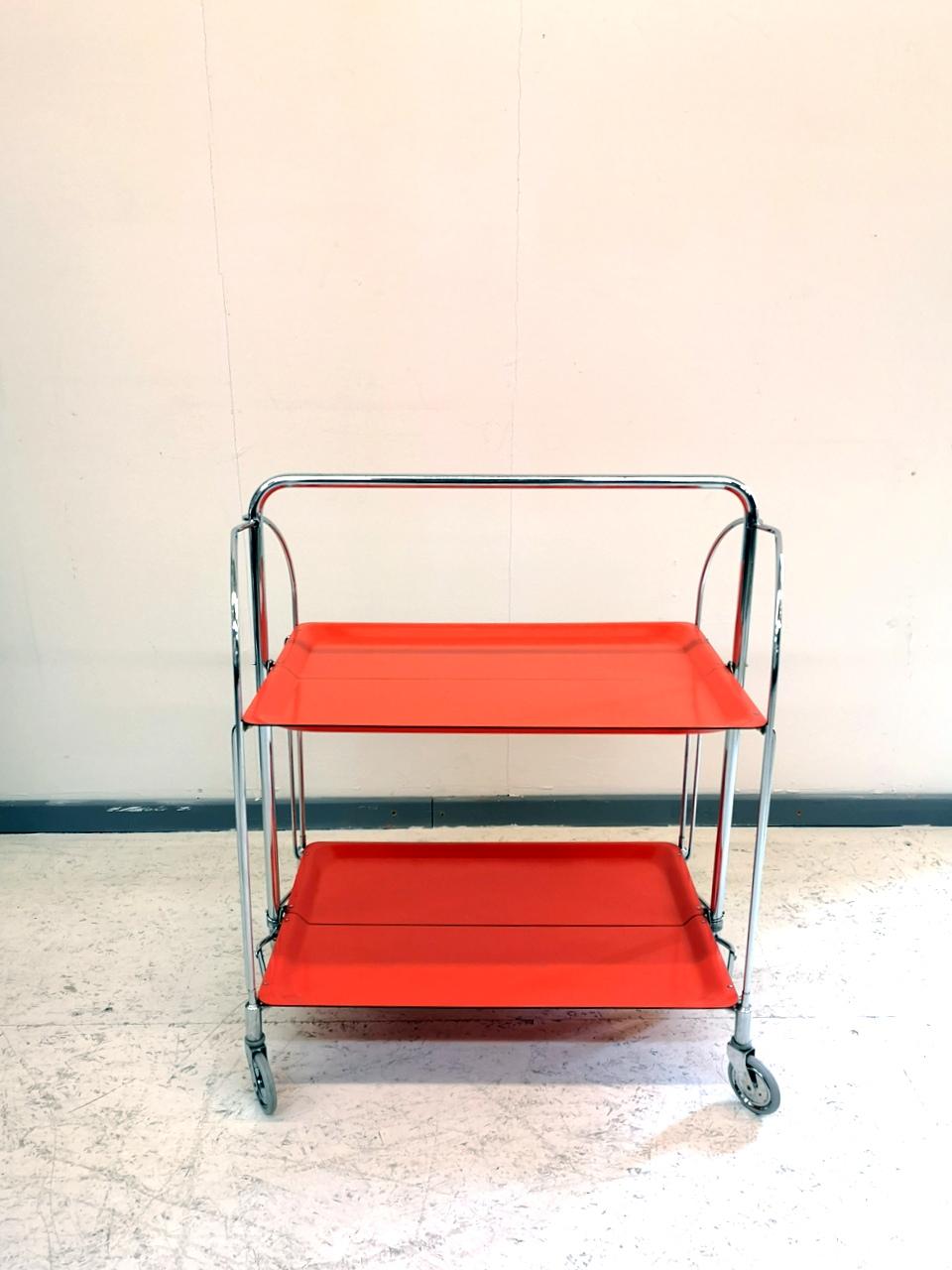 From West-Germany, this 1960s bar cart is foldable in the middle for easy storage. It features a coral/orange color laminated plastic body and chrome-plated legs.