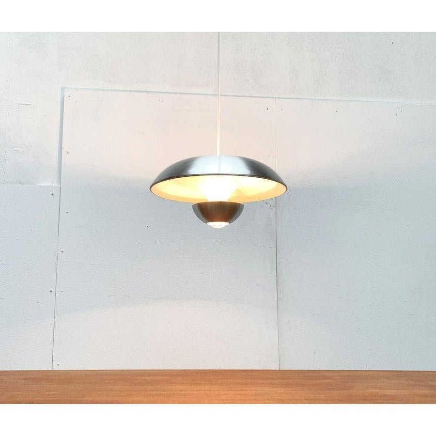 Rara lampada a sospensione dell'era spaziale di Kaiser glow.

Il design colpisce per la sua complessa forma a ombrello con una combinazione di elementi concavi e convessi. Il riflettore bianco sul lato inferiore è realizzato a tulipano e