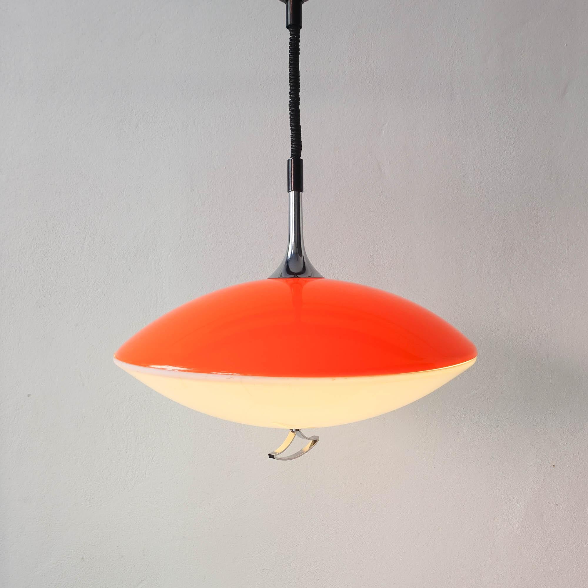 This Space Age UFO pendant lamp was designed and produced in Italy during the 1970's. It has two acrylics, one in orange and other in white, that gives a warm light to the space. With rise and fall mechanism in good condition and a silver pull on