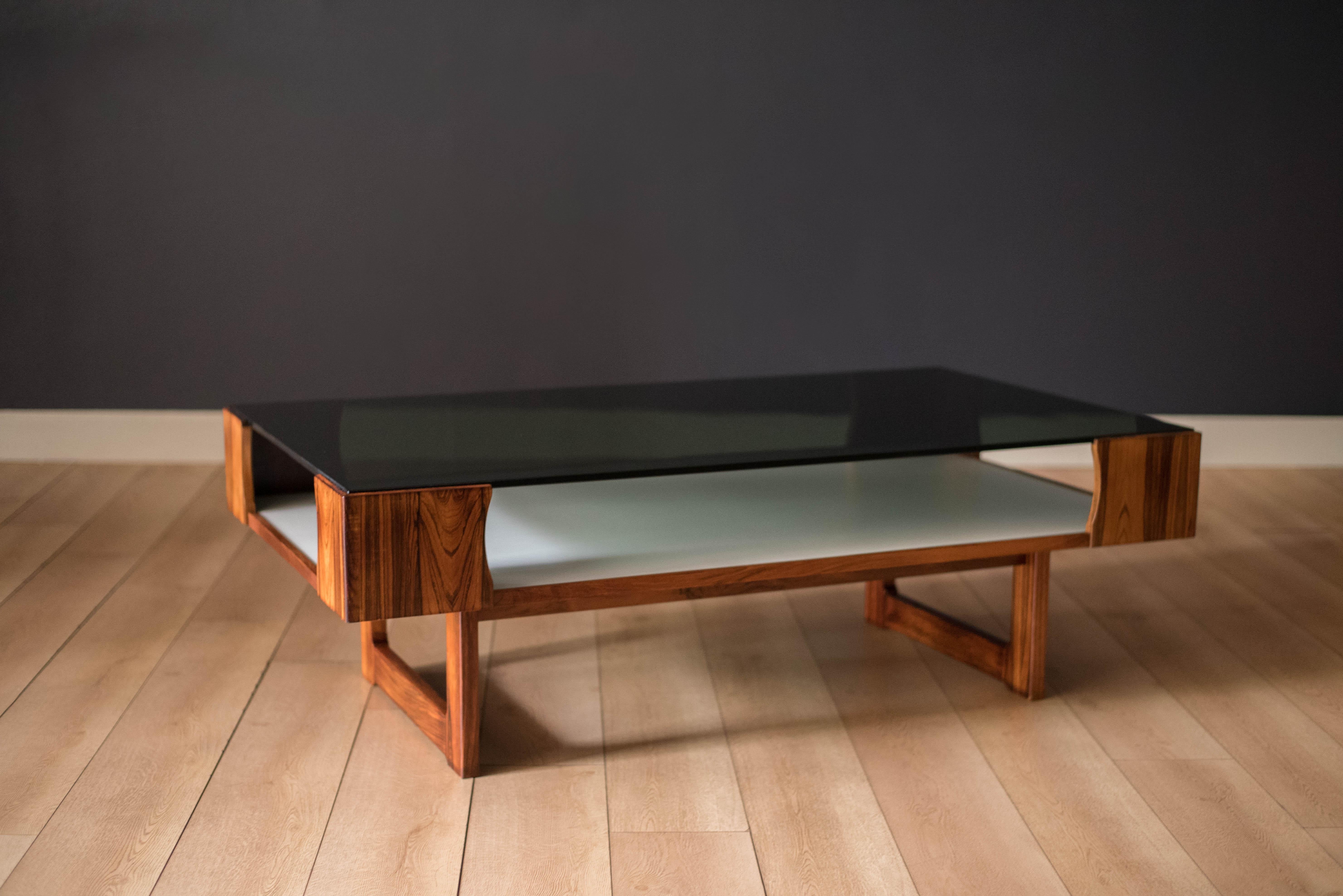 Vintage two tier coffee table in rosewood designed by Torbjorn Afdal for Bruksbo, Norway c. 1970's. This piece showcases from all angles featuring a smoked glass table top with a lower magazine shelf in white laminate for extra storage and easy