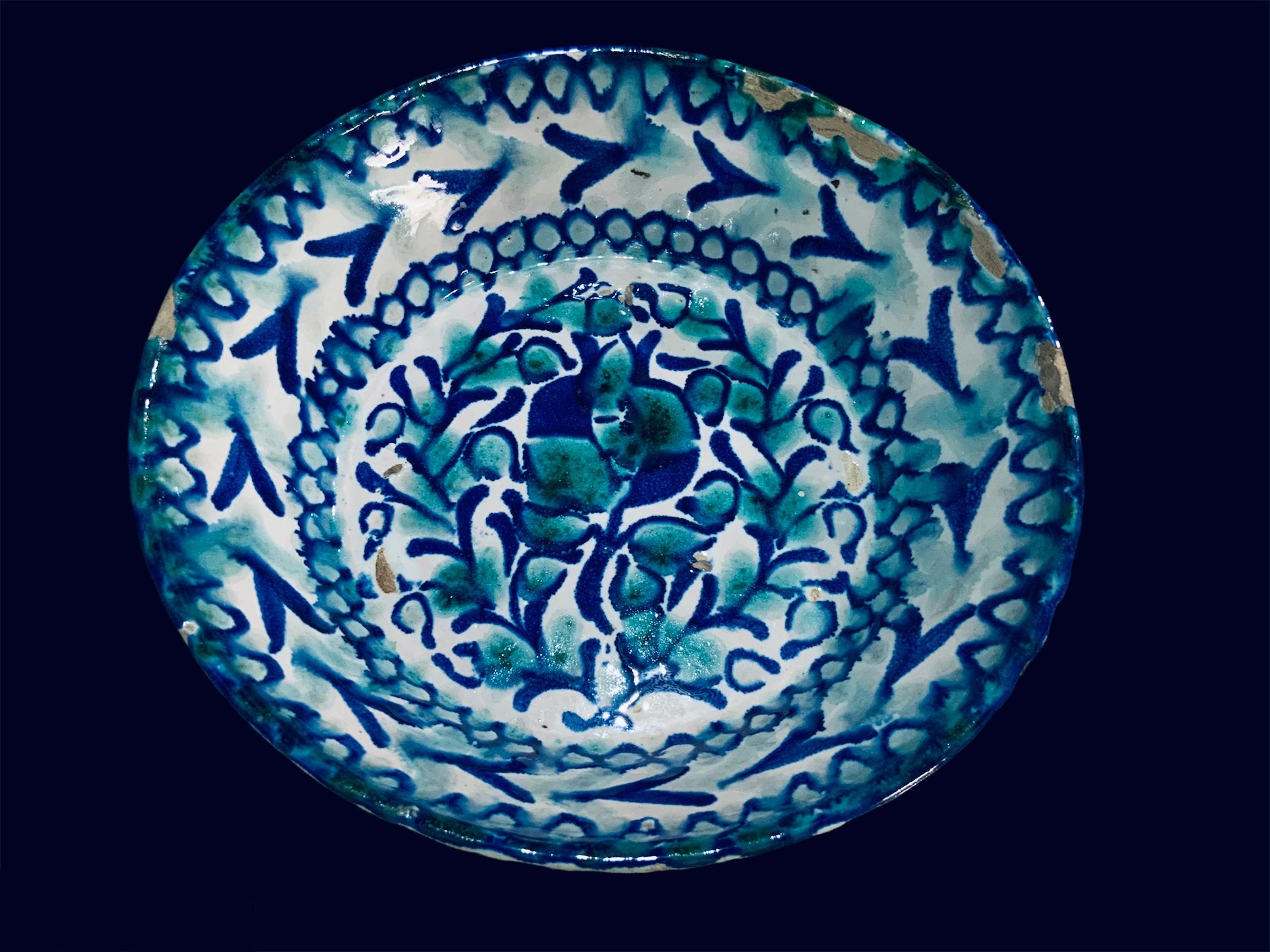 This is a Fajalauza style ceramic bowl from Spain. It depicts a round bowl hand painted cobalt blue and green with a pomegranate flower and some branches in the center. Around this center, there is a garland of circles and above them, some goose