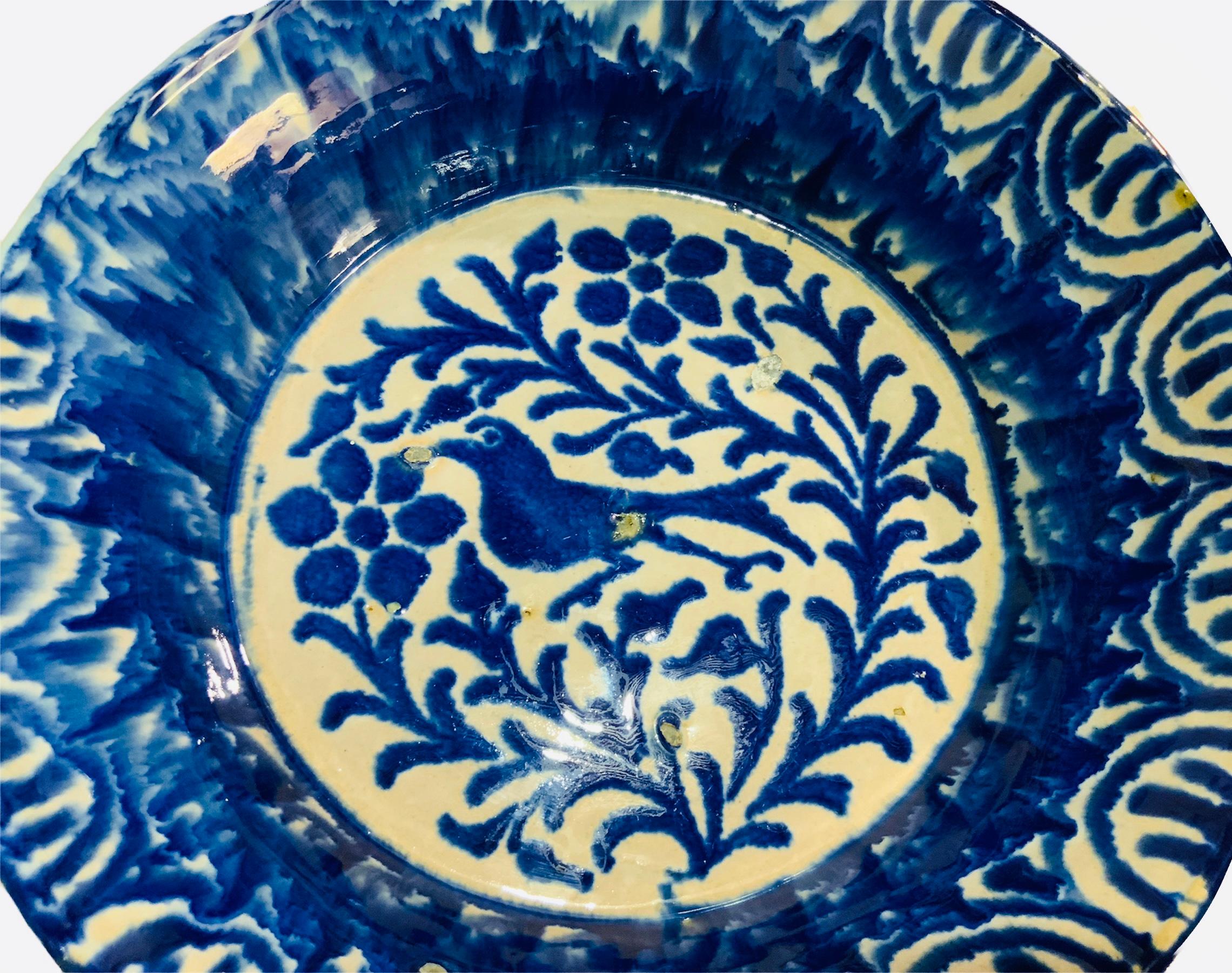 This is a Fajalauza style ceramic bowl from Spain. It depicts a round bowl hand painted cobalt blue and white with a bird and some branches of flowers in the center. Above this center, there is a pattern of cobalt blue splashes of painting. The