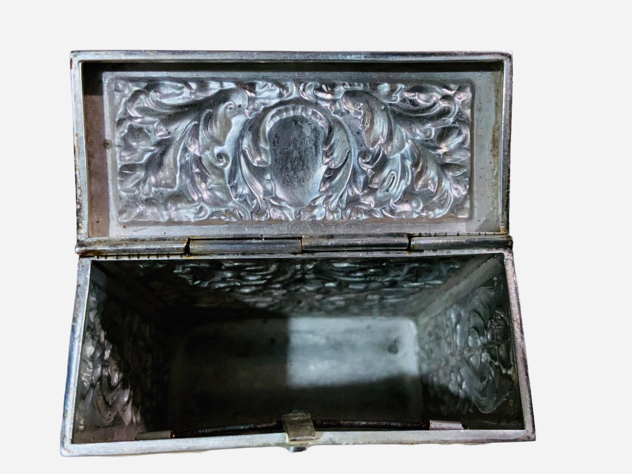 This is a Spaniard Silver Box. It depicts a hinged lid rectangular box adorned with a delicate repousse pattern of acanthus leaves and flowers in the front, back and sides of it. A monogram of three letters- ERO- are inscribed in the top of the lid.