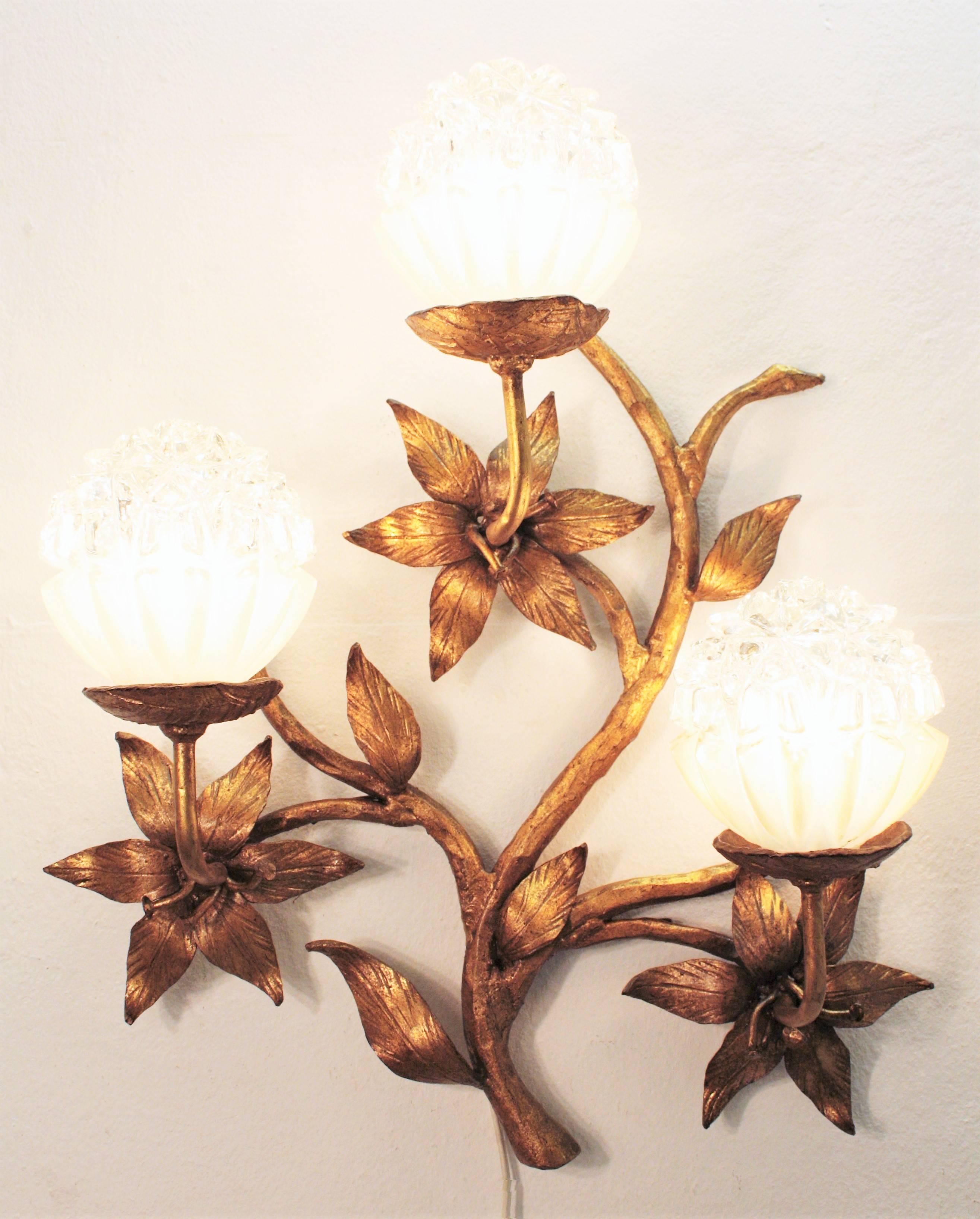 An spectacular hand-hammered iron floral wall sconce with gold leaf gilt. Three branches with a big flower decorating each of them and geometric molded glass globes as lamp shades at the top of each branch. A finely handmade work full of movement