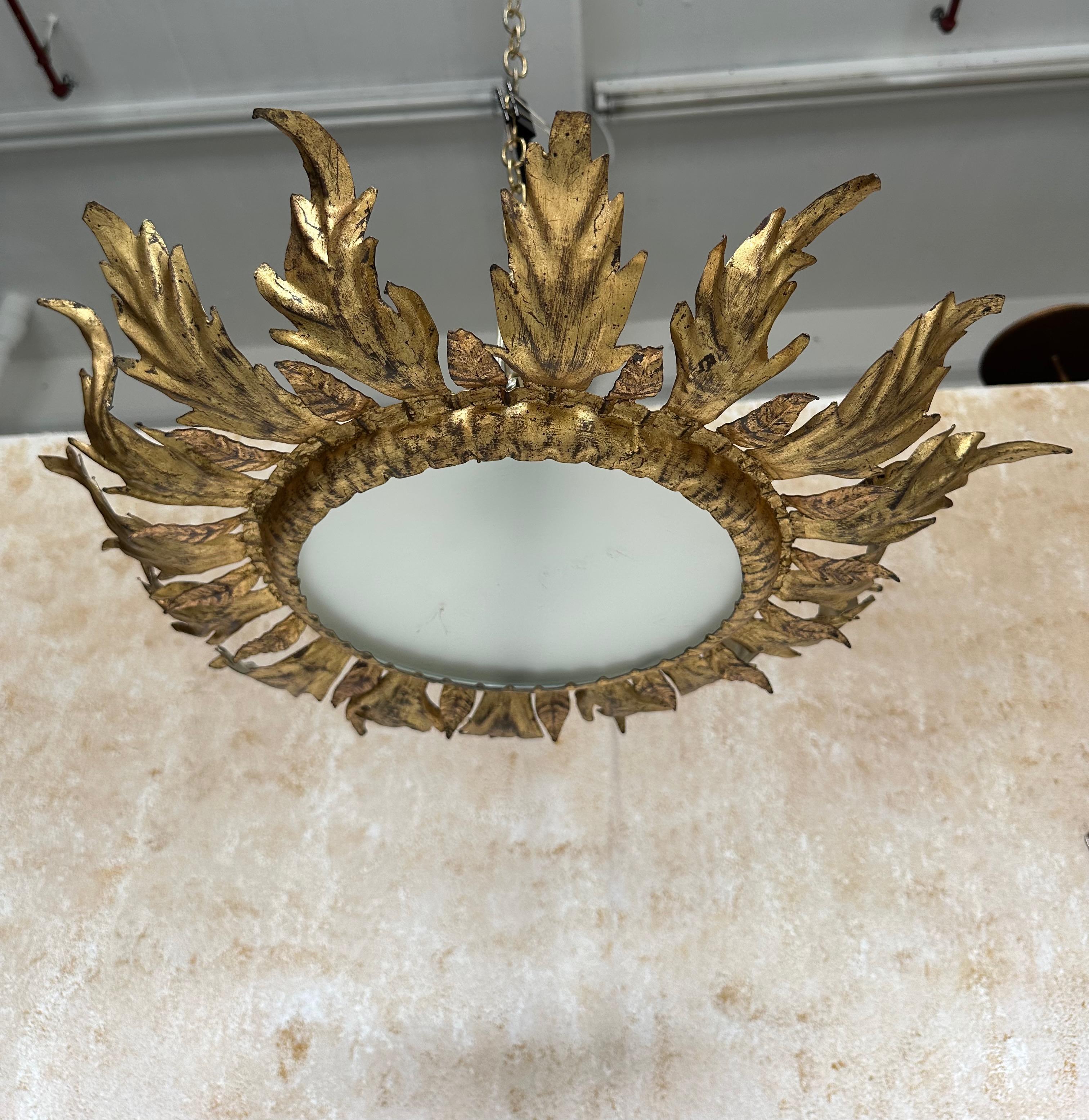 This lovely vintage 1950s Spanish gilt metal ceiling fixture features alternating large and small stylized leaves that curl gently upward surrounding a hammered central ring with a frosted glass center. The rich hand applied gilt finish is original